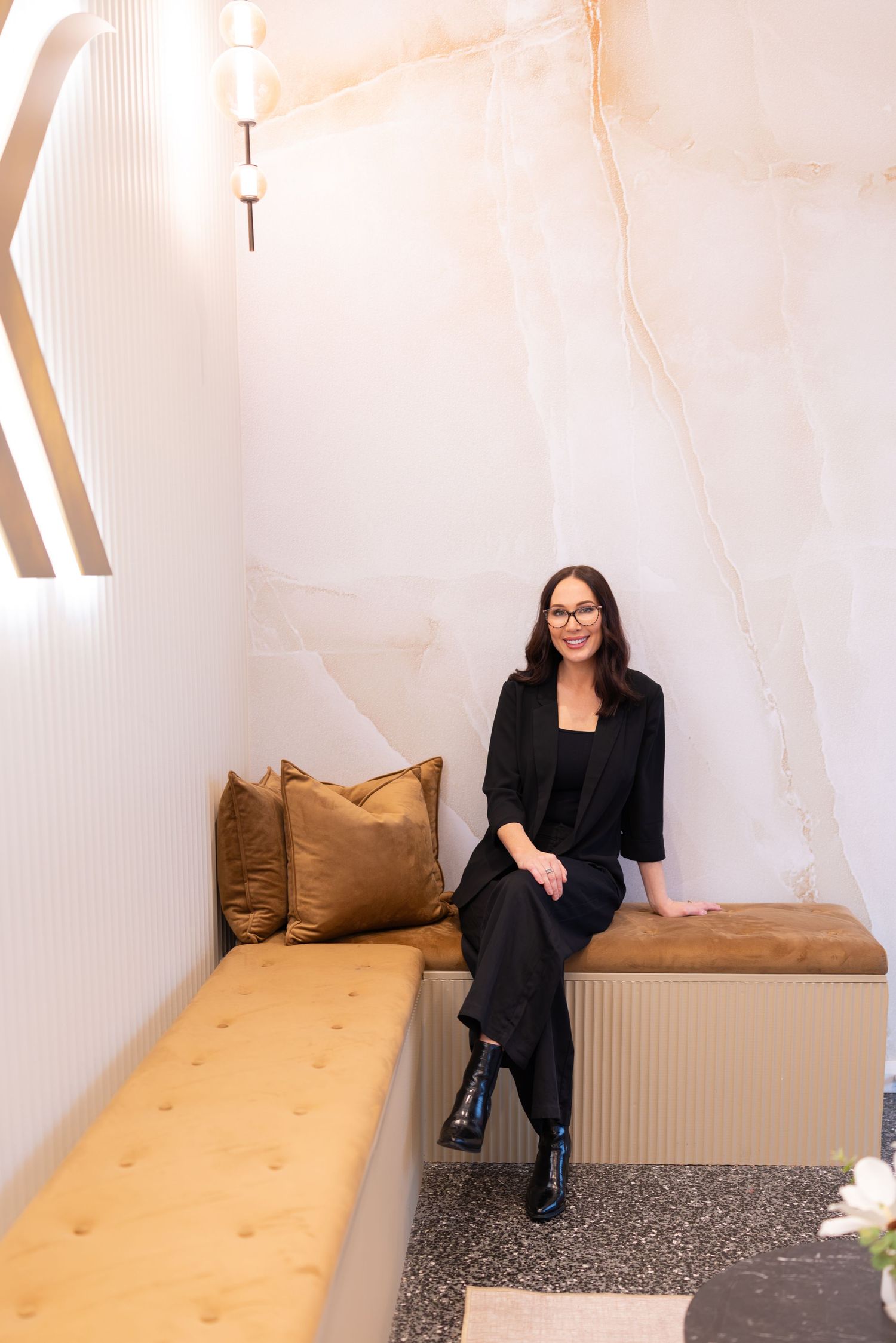 Woman in black outfit sitting on a tufted gold bench with velvet pillows against a marble wall.