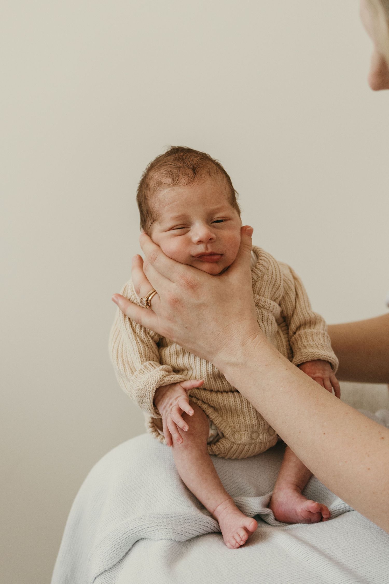 Newborn baby in beige sweater smiles while being held on white background.