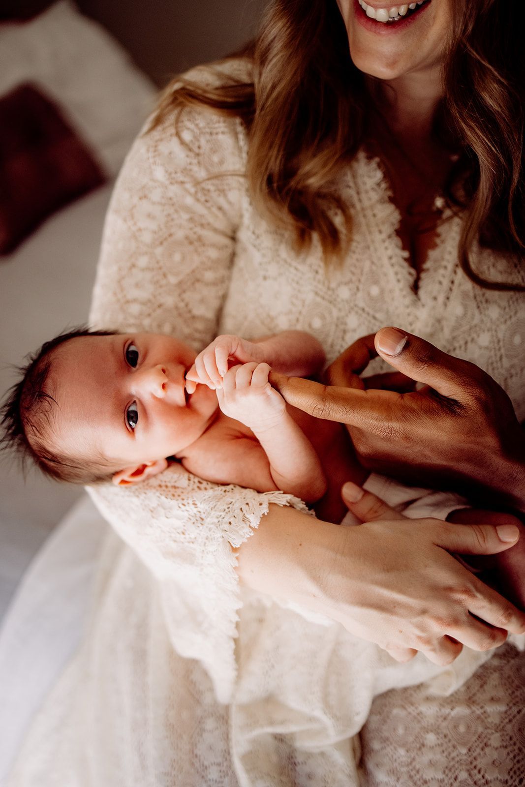 Smiling mother in white lace dress cradling her newborn baby, who gazes up with wide eyes while sucking fingers.