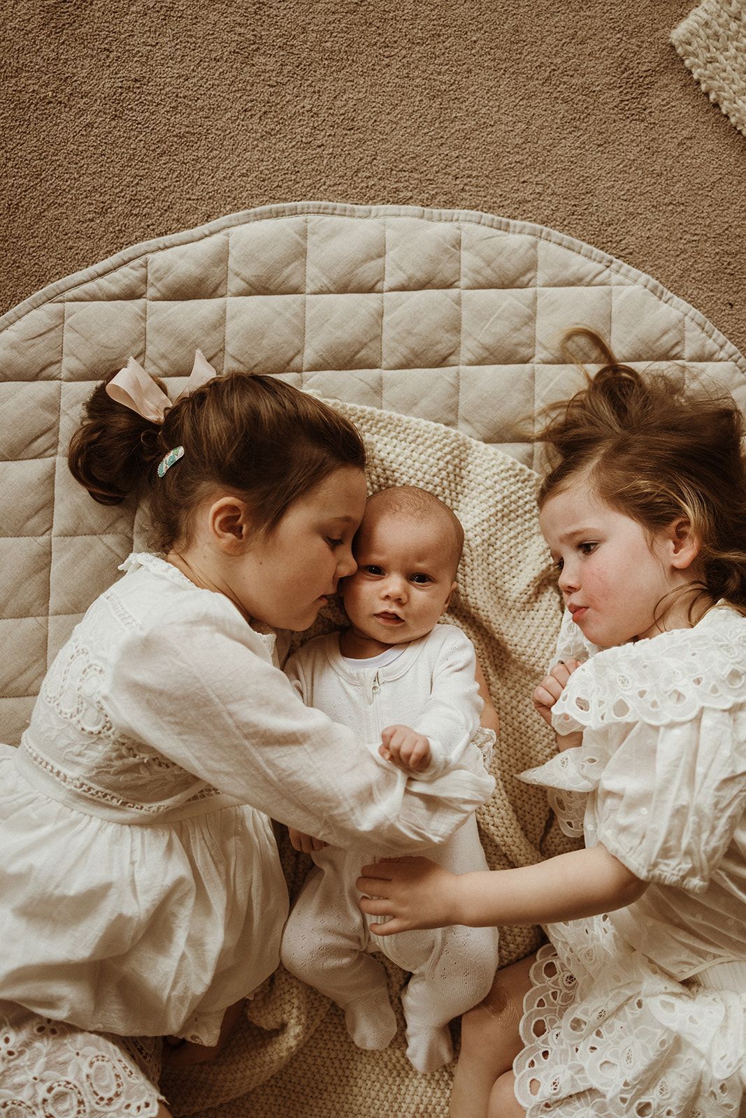 Two young girls in white dresses lovingly cuddle a newborn baby on a quilted beige play mat.