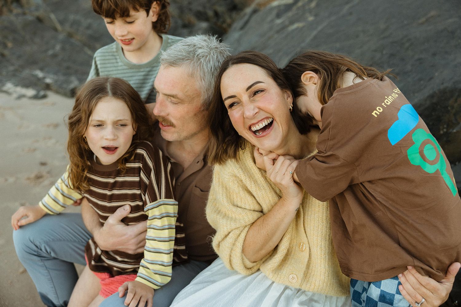 A joyful family of five laughing together outdoors near rocky cliffs, with parents and three children sharing a warm embrace.