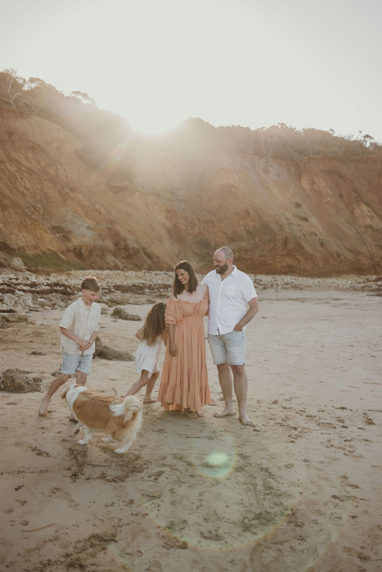 Family with two kids and a dog playing on a sandy beach at golden hour with cliffs in the background.