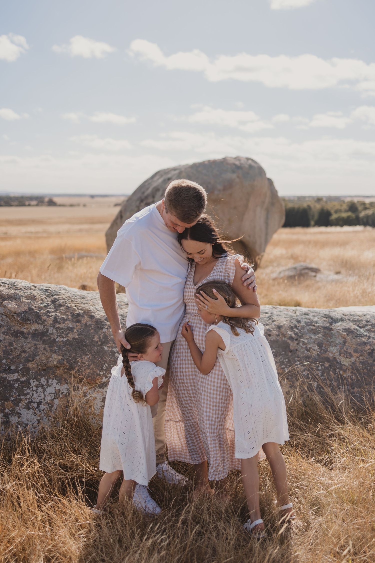 Family of four embracing outdoors in golden field, parents in white and checkered outfits with two young girls in white dresses.