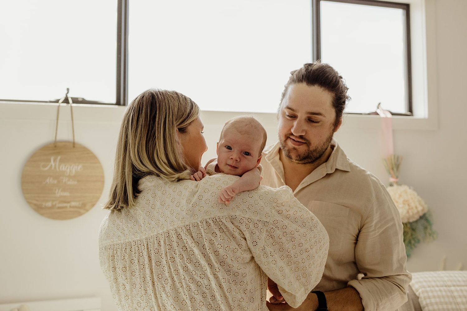 A couple admires their newborn baby in a bright, cozy room with a personalized wooden name sign on the wall.