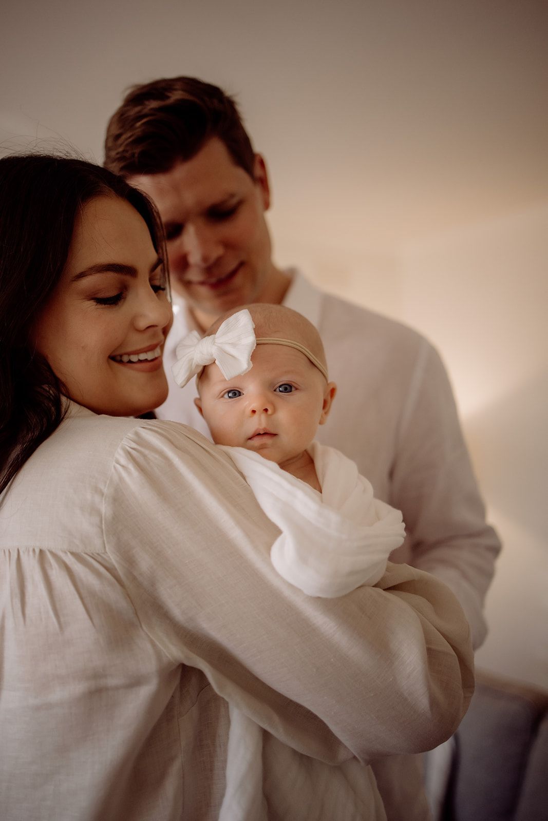 Happy young parents in white clothing lovingly holding their newborn baby girl with a white bow headband.