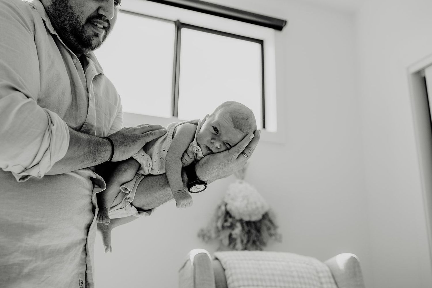 Black and white photo of a father lovingly holding his newborn baby near a bright window indoors.