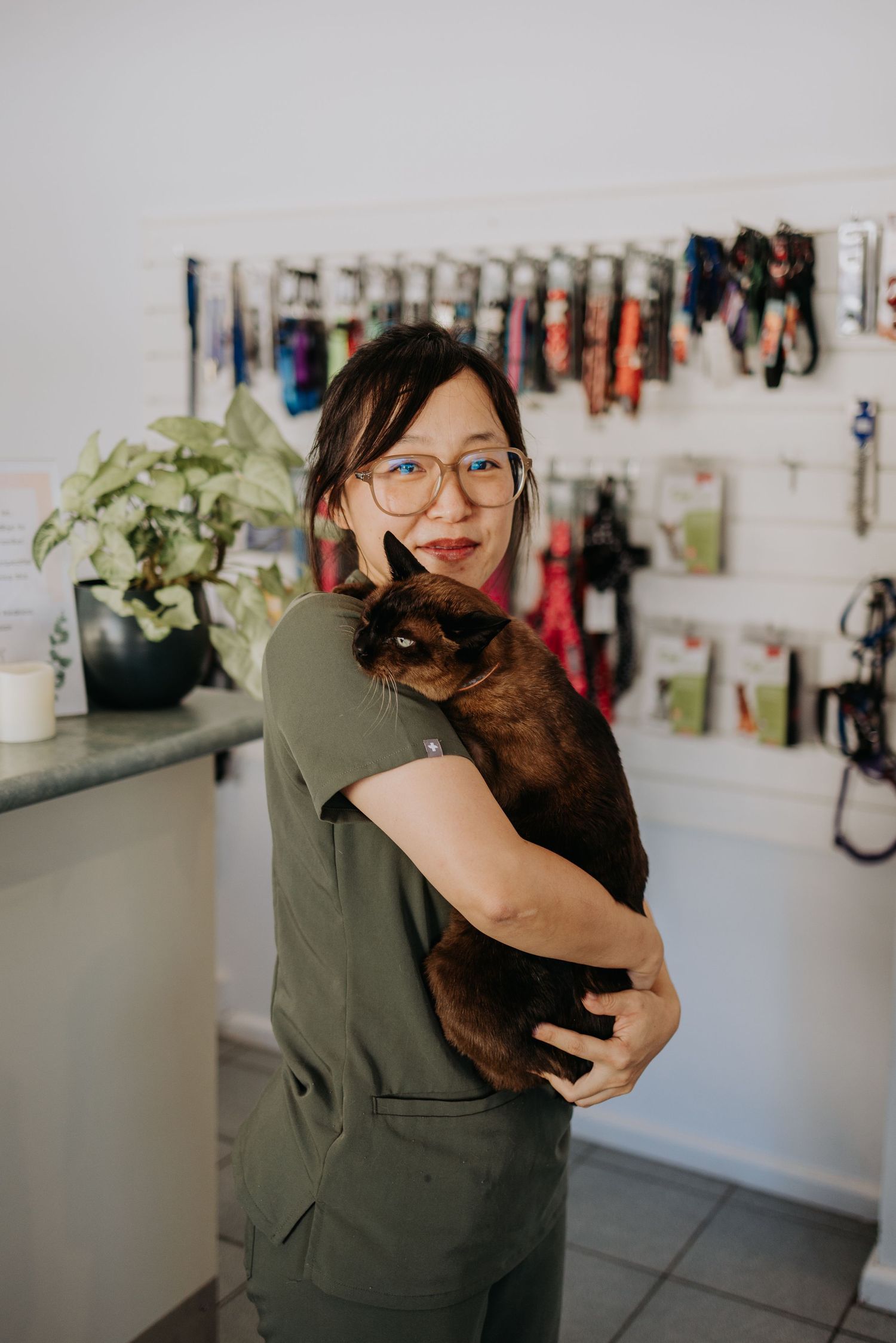 A smiling woman in olive scrubs holds a brown Burmese cat in a veterinary clinic with leashes on the wall.