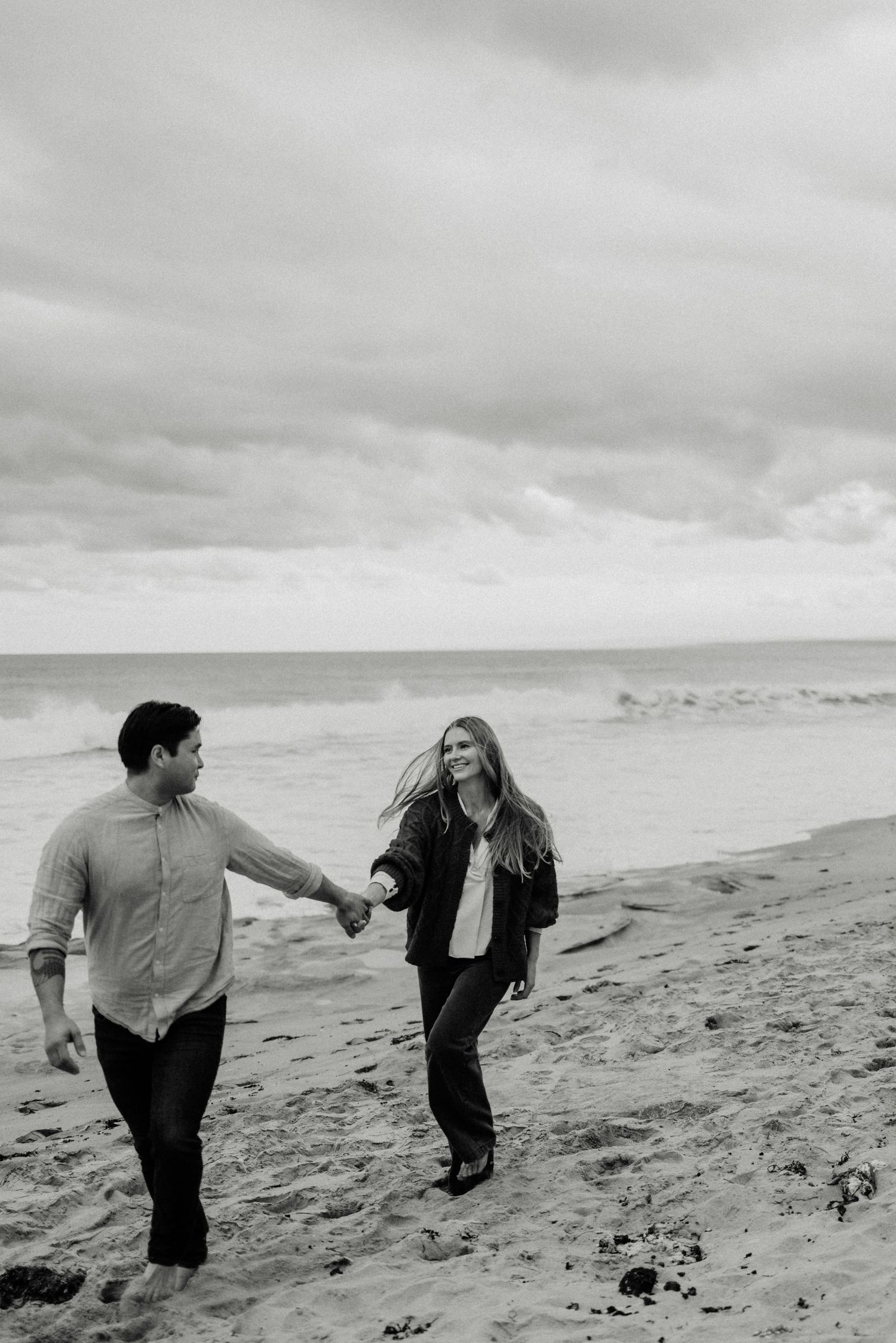 Black and white photo of a couple holding hands and walking on a sandy beach with ocean waves in the background.