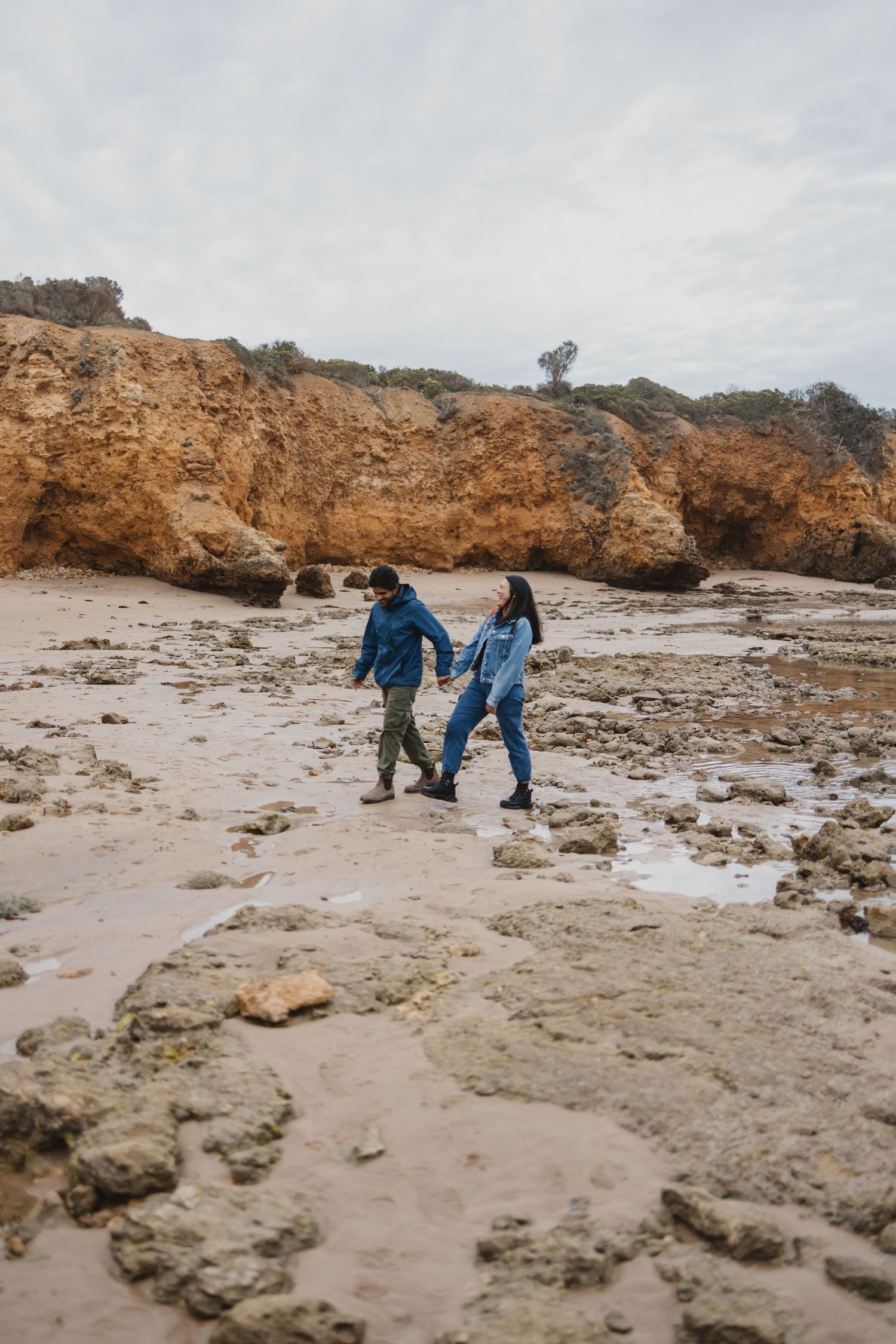 Couple holding hands walking on a rocky beach with golden cliffs in the background on a cloudy day.