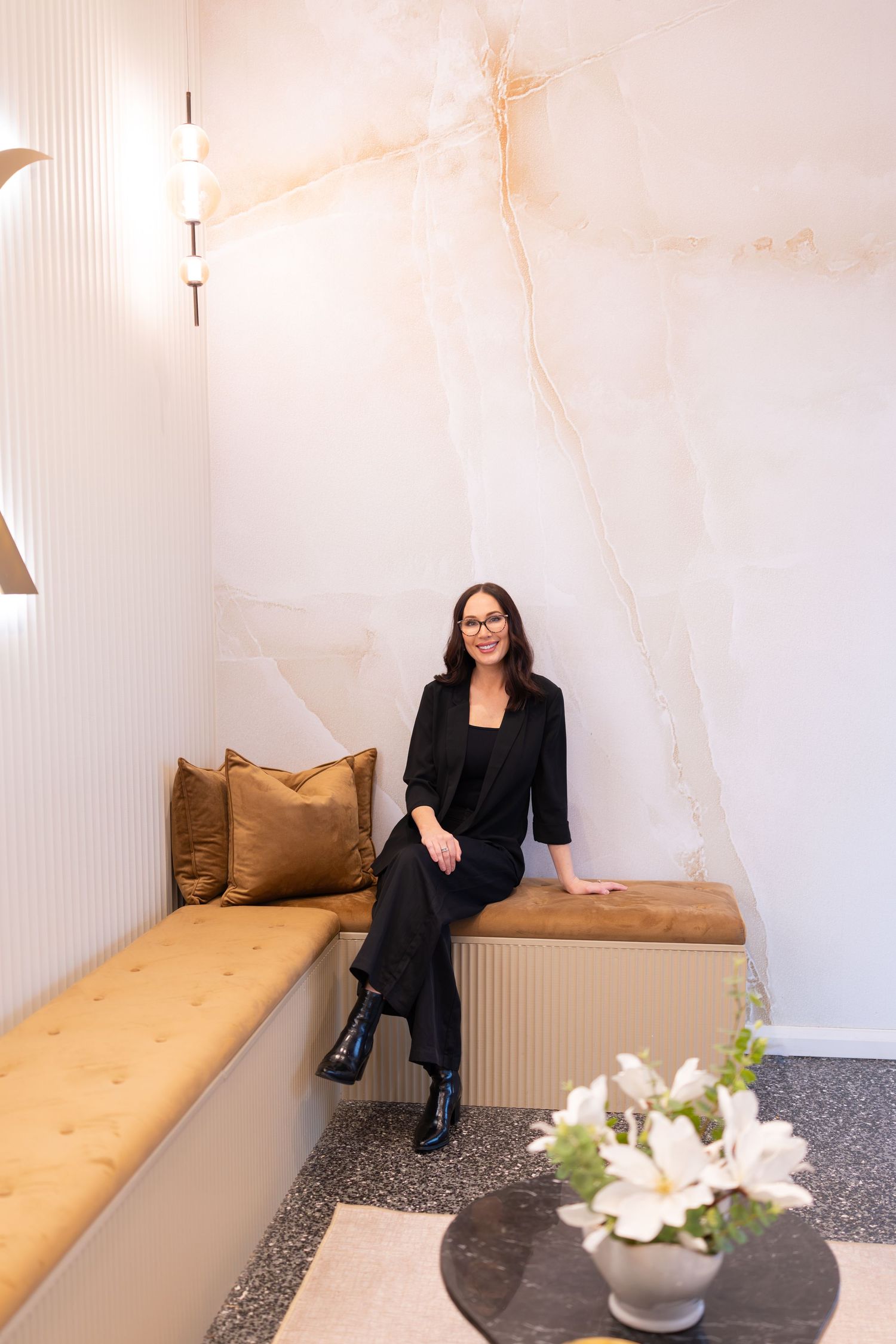 Woman in black outfit sitting on gold bench against marble wall in elegant interior space.