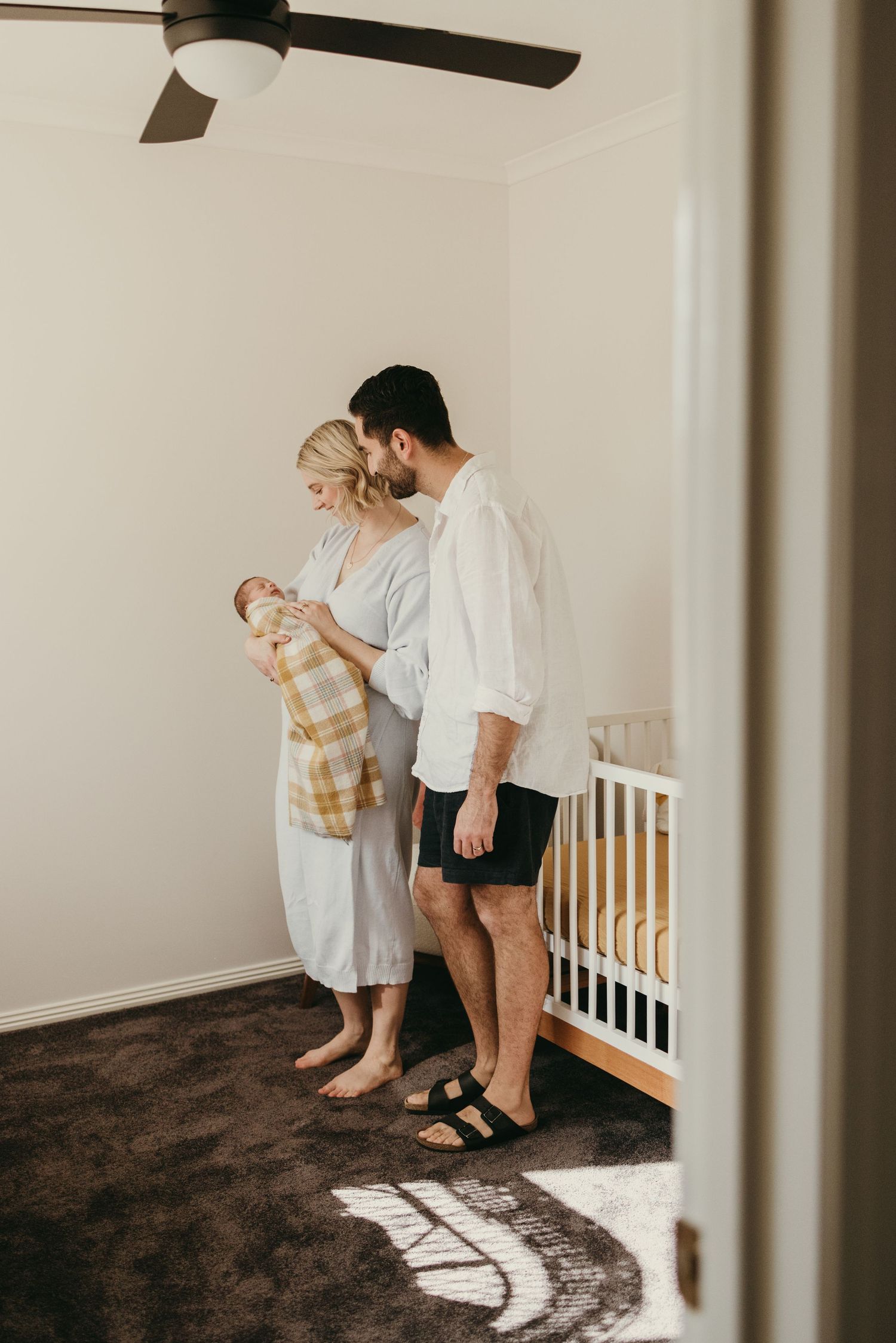 New parents stand together in nursery with ceiling fan looking down at their swaddled baby.