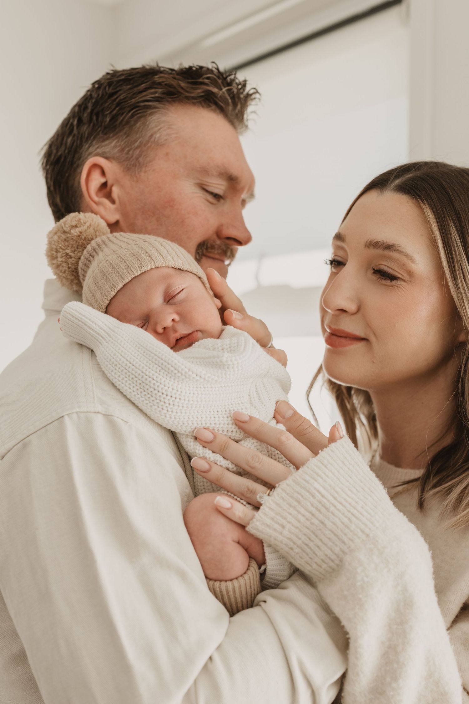 A couple tenderly holds their sleeping newborn baby dressed in a white knit outfit and beige bear hat.