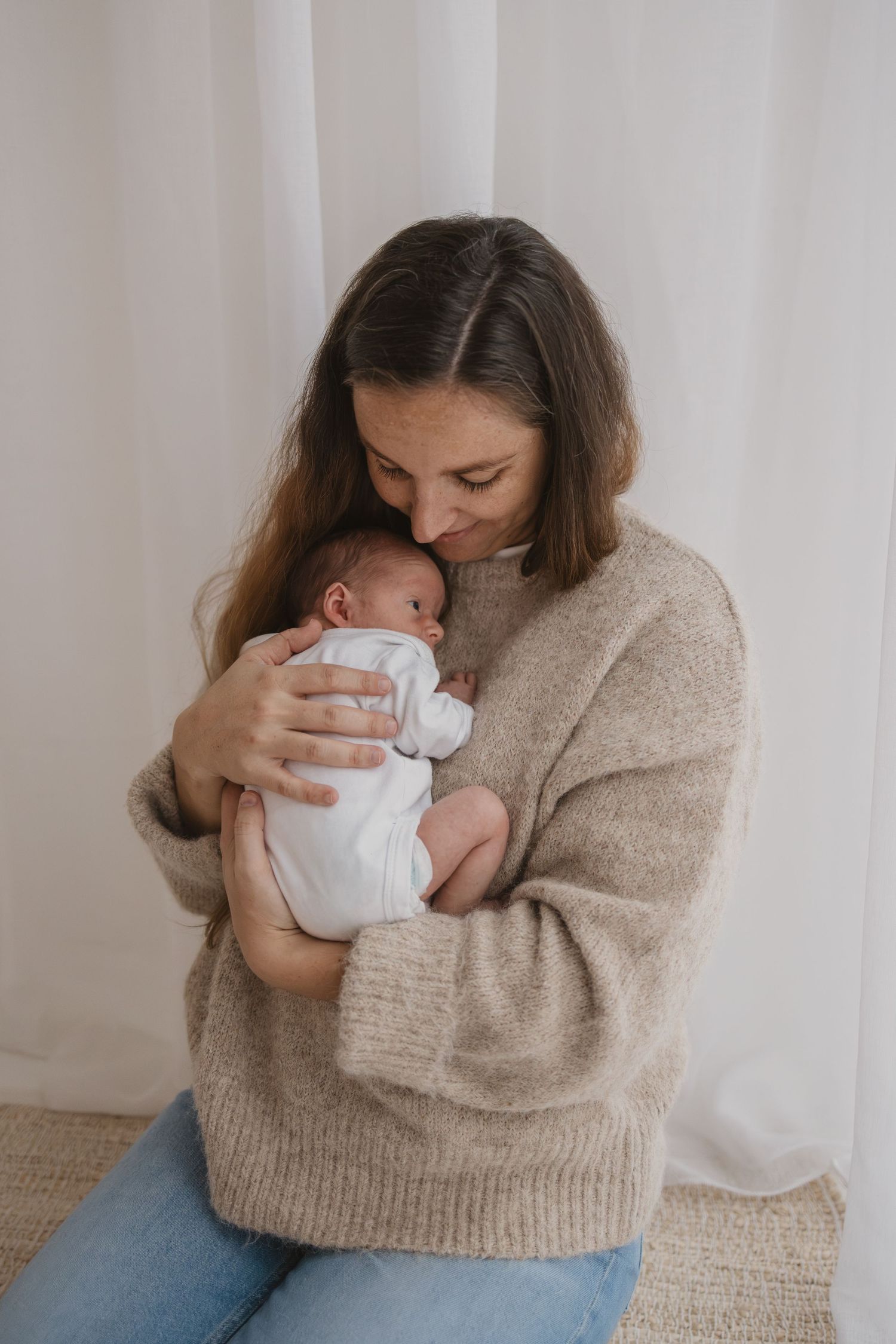 A mother in a beige sweater tenderly holds and kisses her newborn baby dressed in white.