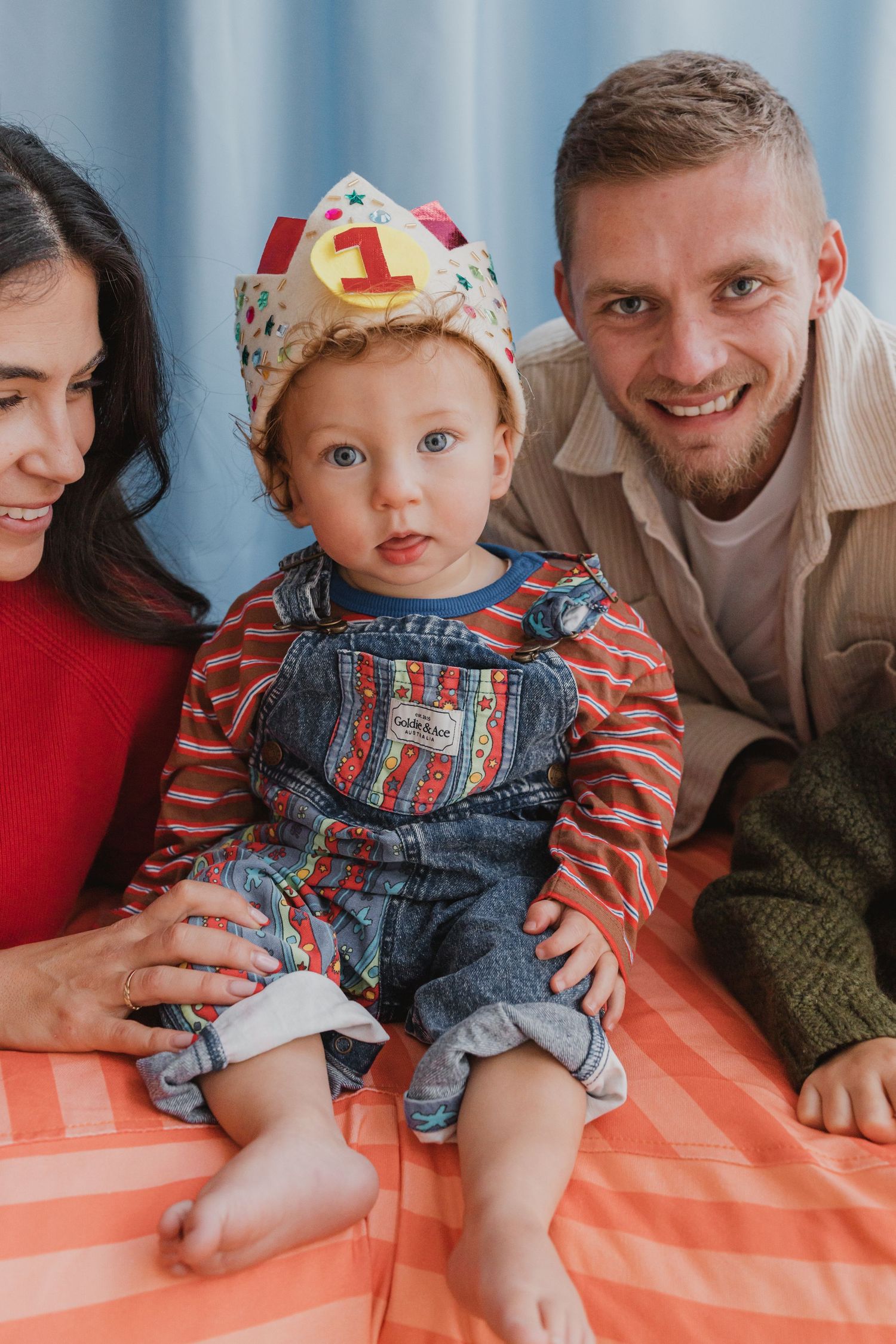 A baby wearing a number 1 crown sits between smiling parents on their first birthday.