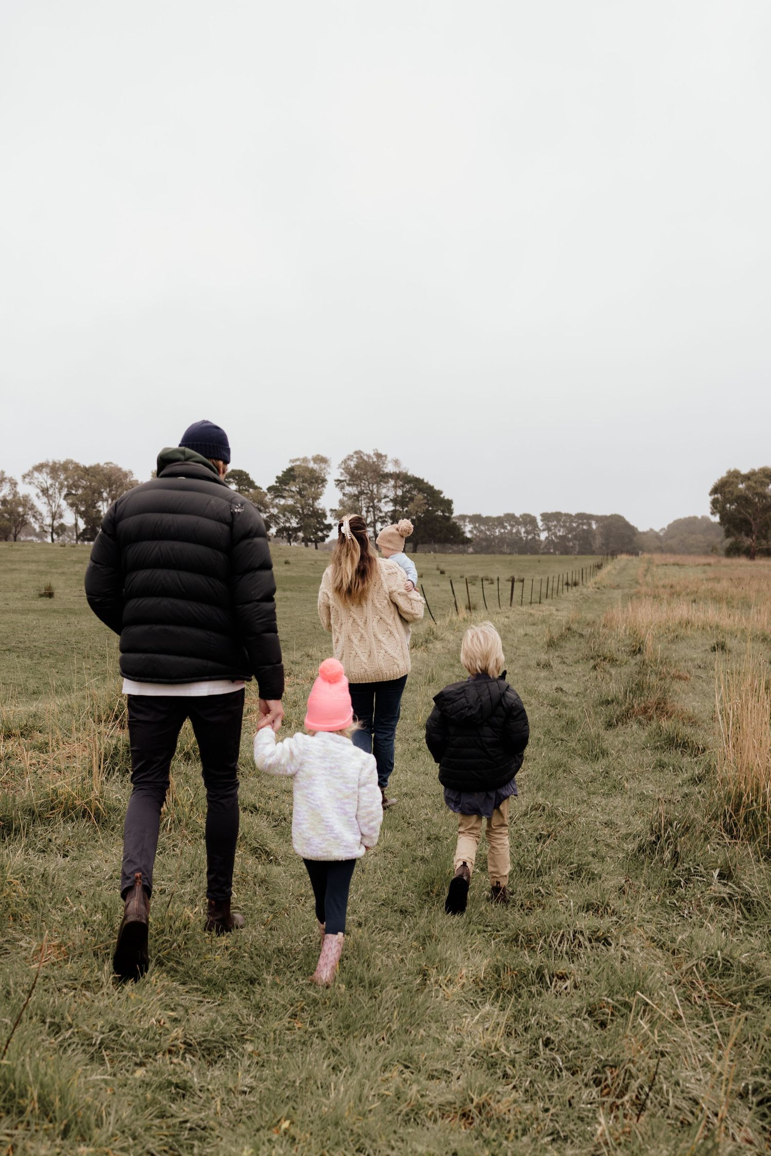 Family of four walking through a green field on a cloudy day, wearing winter jackets and holding hands.