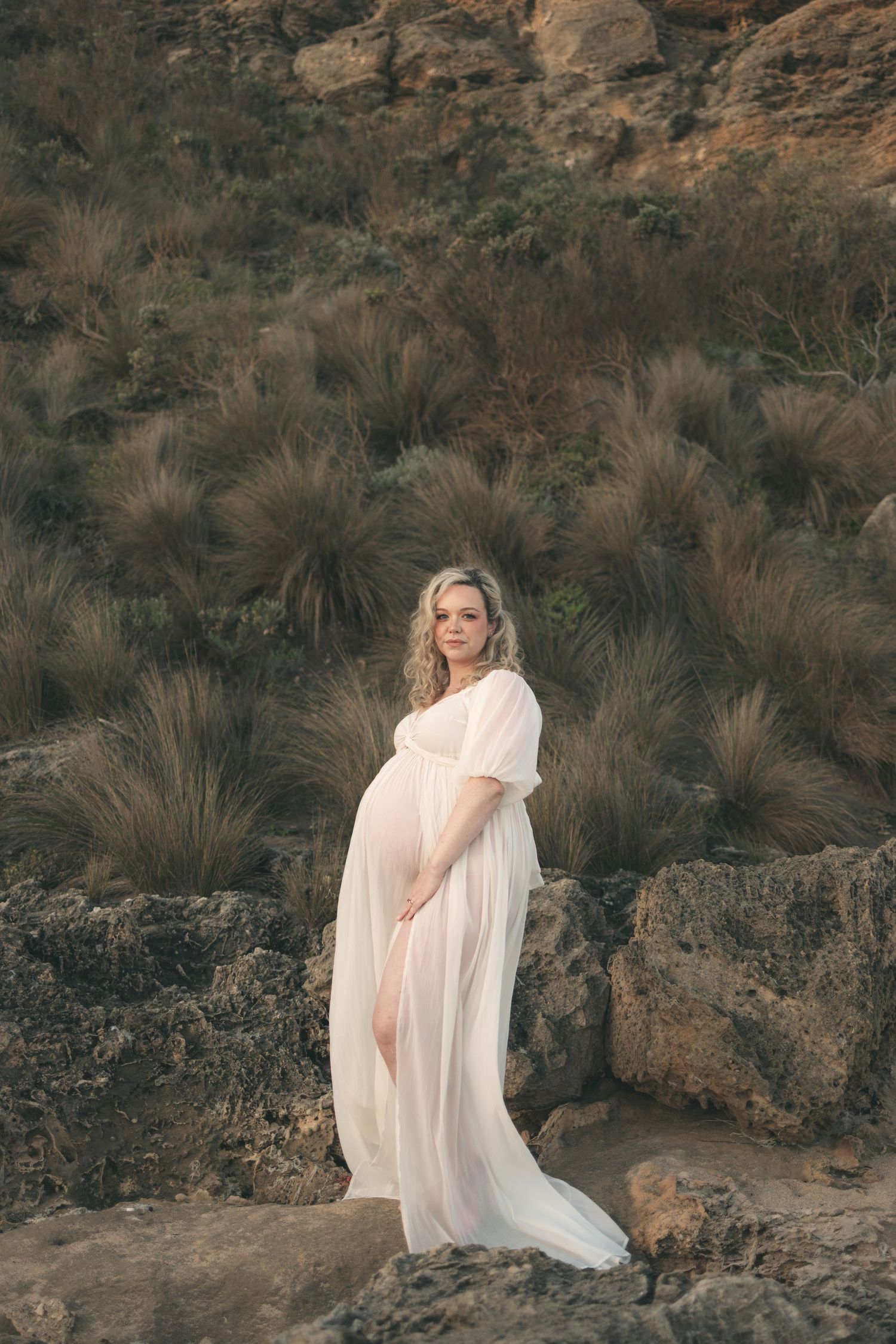 Pregnant woman in sheer white maternity gown poses on rocky terrain with desert shrubs in background.