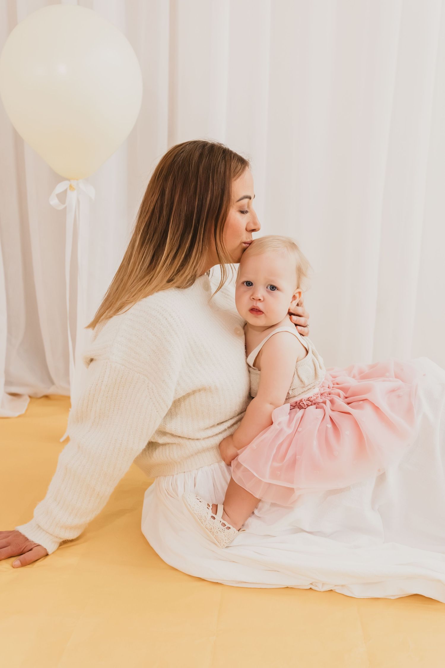 A mother in a white sweater kisses her baby girl in a pink tutu dress during a birthday photoshoot.