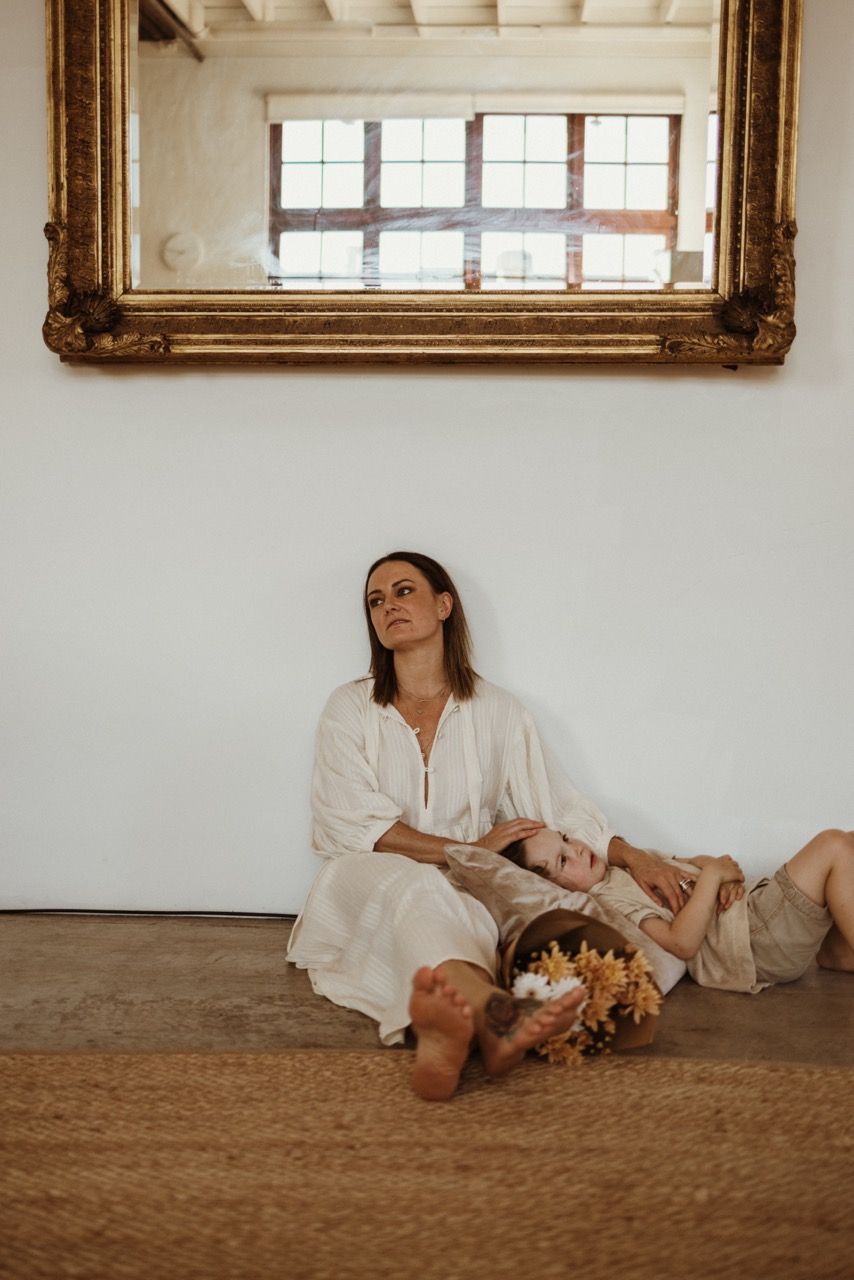 Figure in white dress sits contemplatively in corner beneath ornate gilt frame mirror in rustic setting.