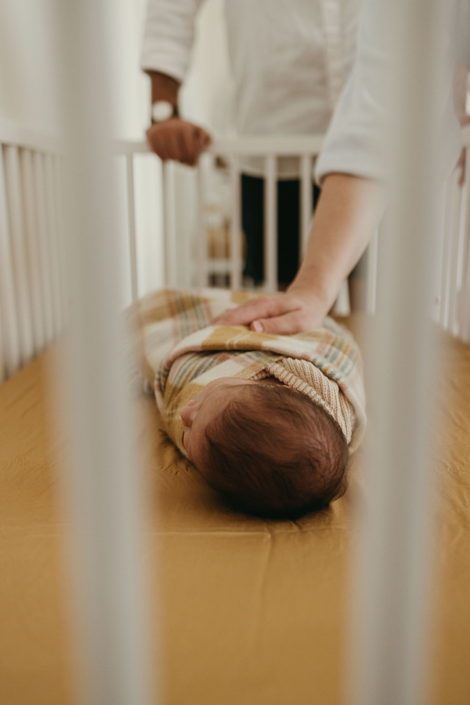 Two hands gently touch a swaddled baby lying on a mustard-colored mattress in a white crib.