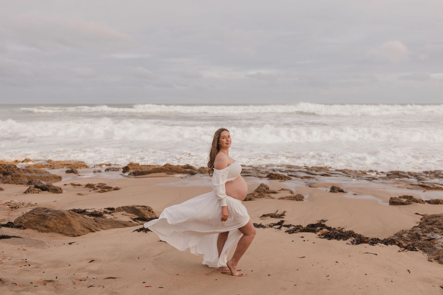 Pregnant woman in white flowing dress poses gracefully on sandy beach with rocky shoreline and ocean waves behind her.
