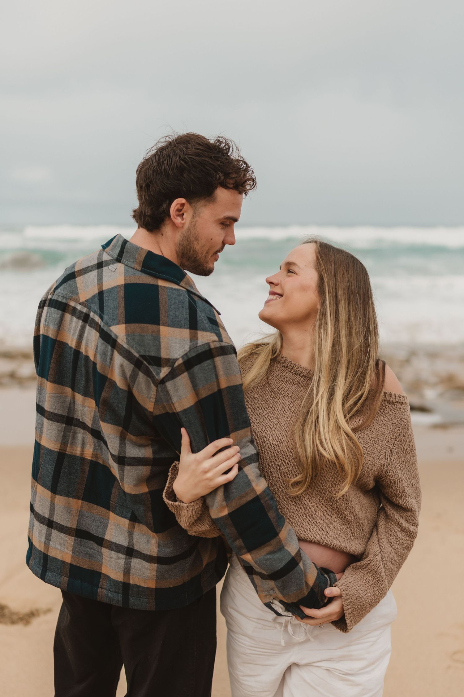 Couple embracing and smiling at each other on a beach, wearing cozy sweaters on a cloudy day.