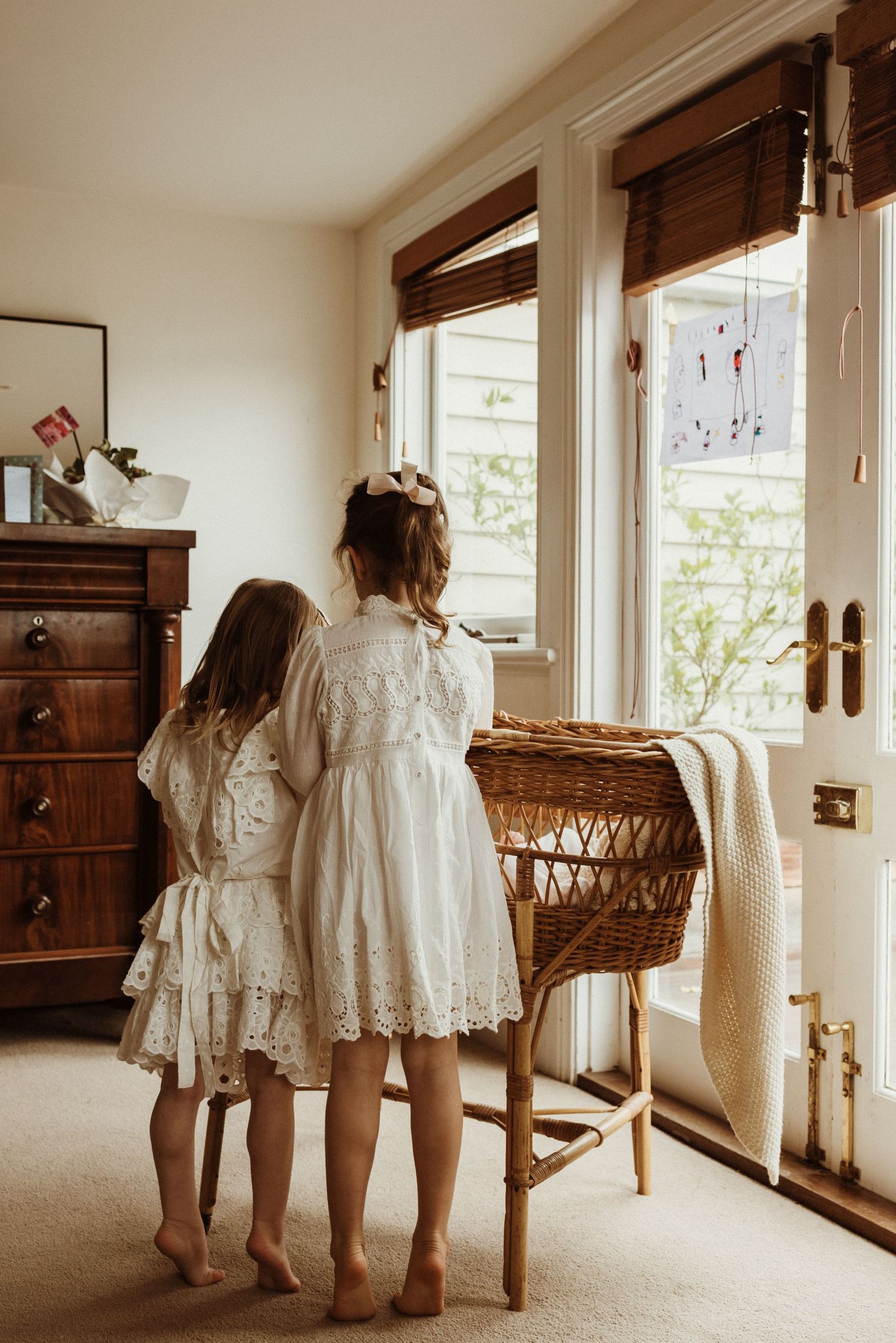 Two young girls in white lace dresses stand barefoot looking out a window beside a wicker bassinet.