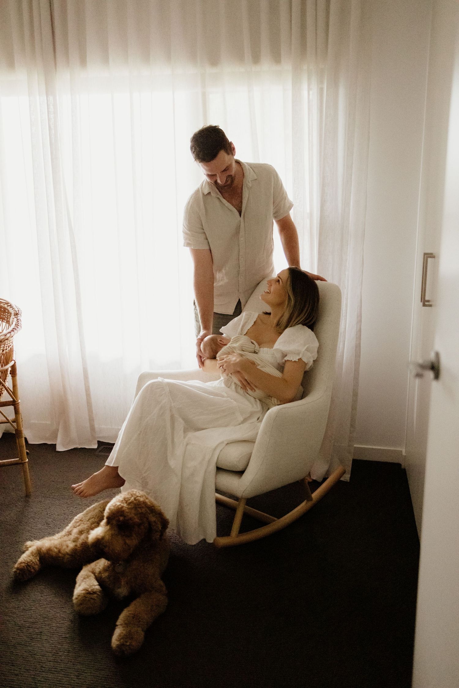 New parents share tender moment, mother in white dress holds newborn in rocking chair beside proud father.