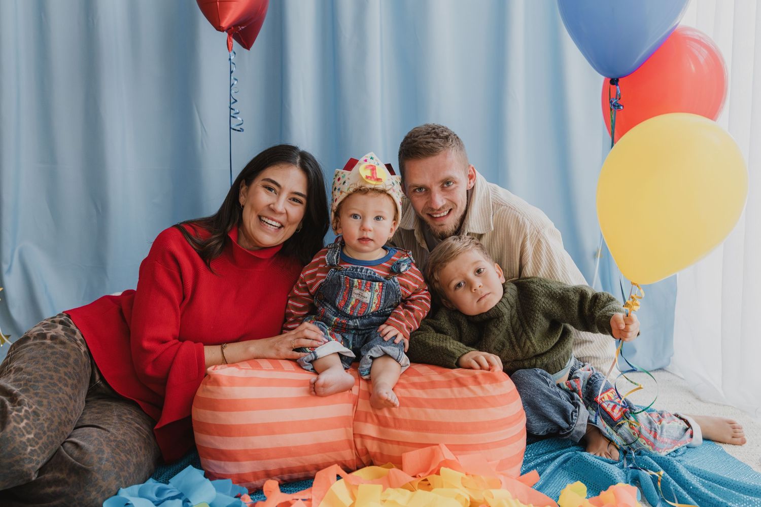 Happy family with two children celebrating birthday with colorful balloons and orange poufs on blue backdrop.