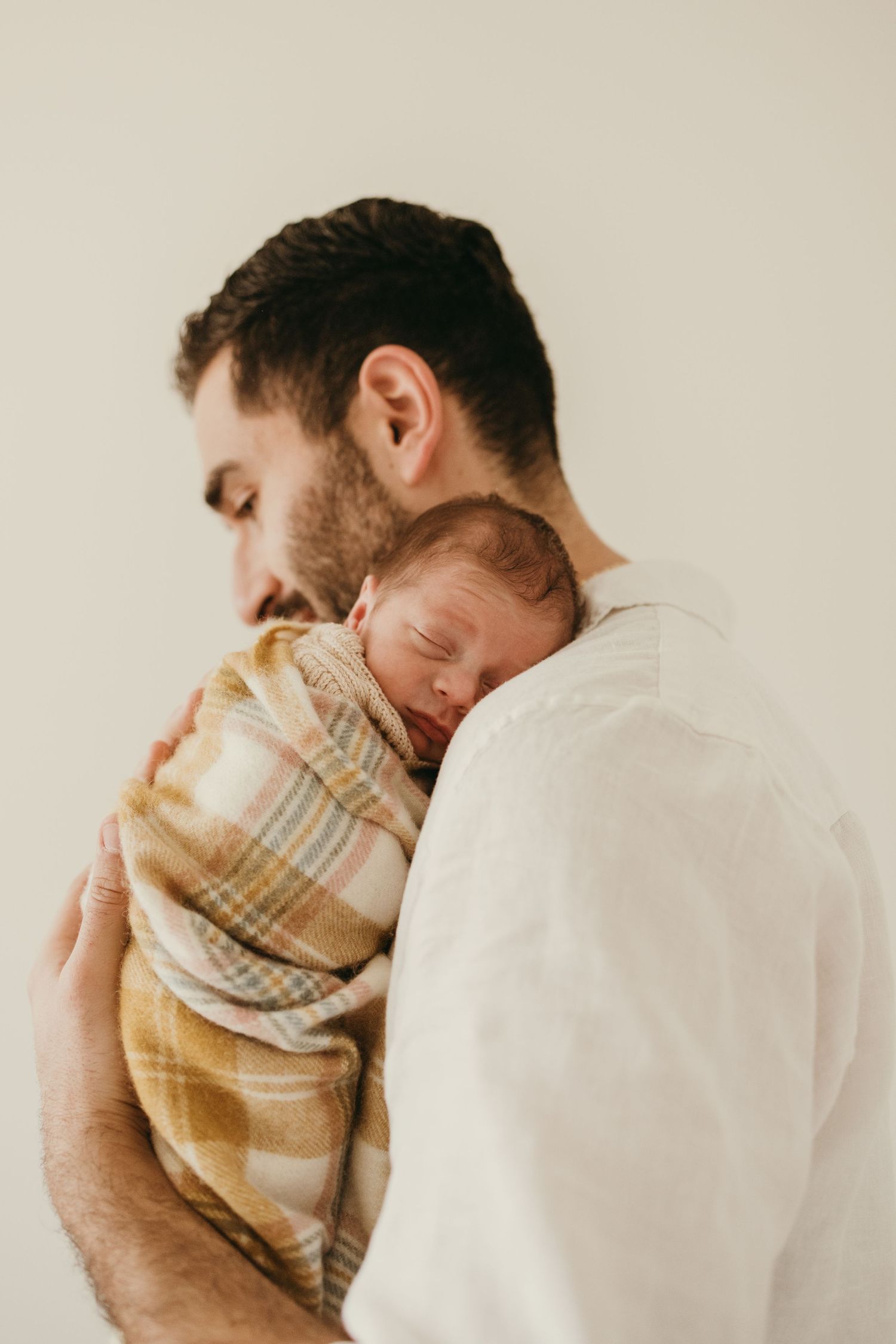 A tender moment between a parent and newborn swaddled in a plaid blanket against a neutral background.