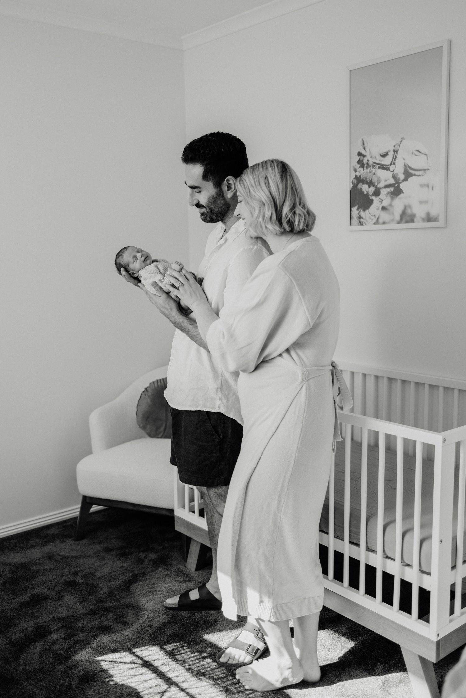 Black and white photo of parents cradling their newborn baby in a minimalist nursery setting.