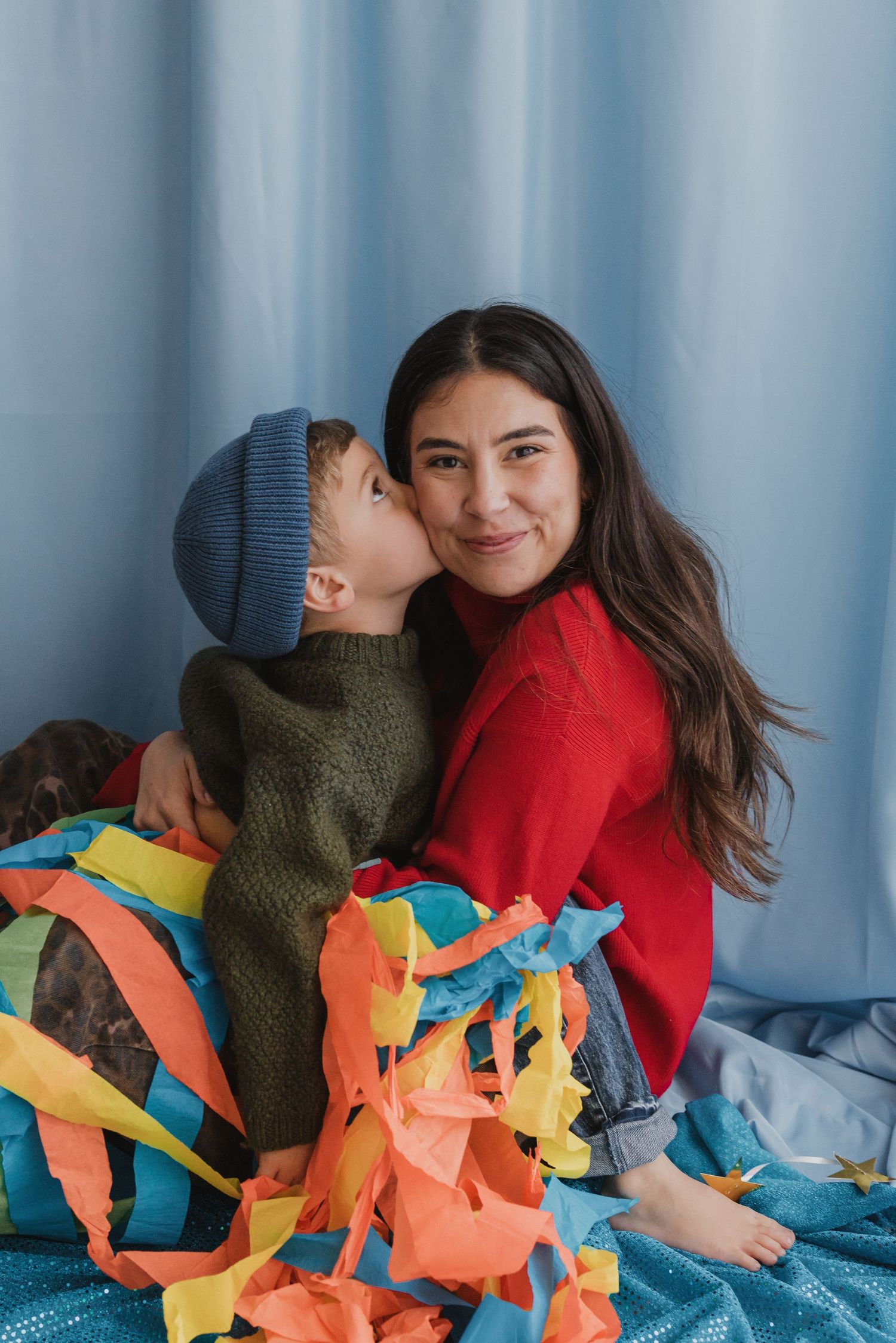 A young boy in a green sweater kisses a smiling woman in red on the cheek, surrounded by colorful paper decorations.