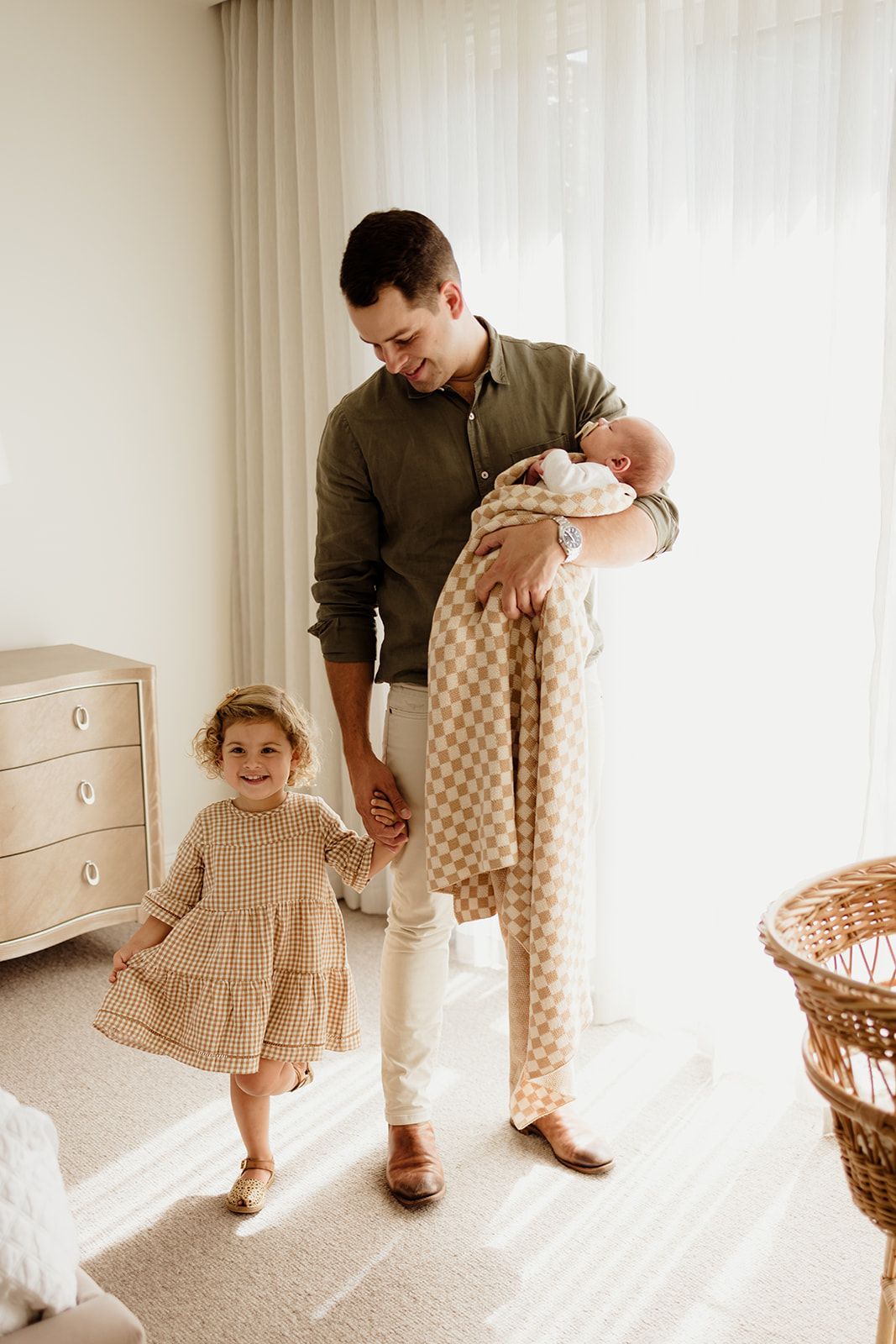 Father holding newborn baby in checkered blanket while toddler girl in tan dress smiles and holds his hand indoors.