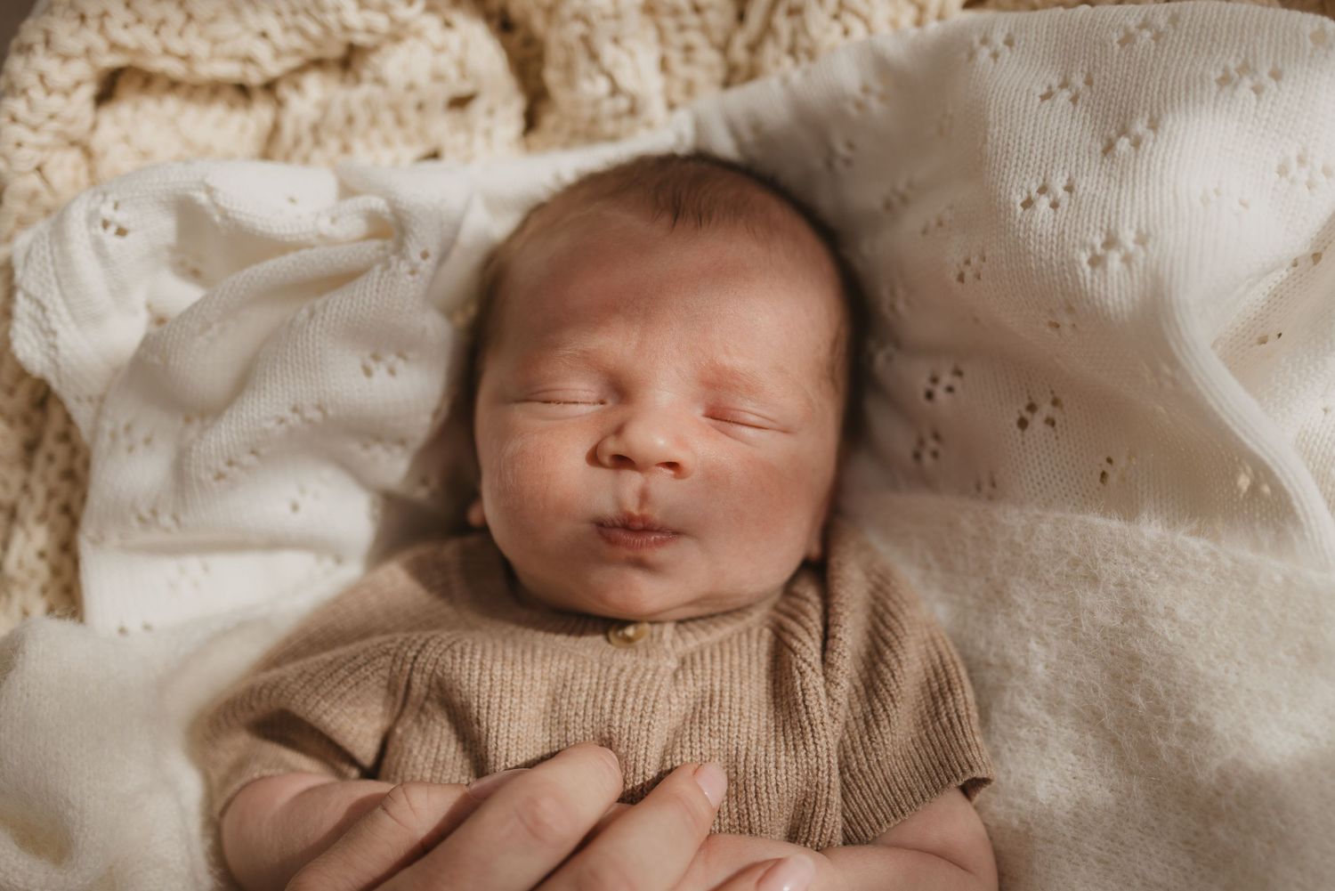 Peaceful newborn baby sleeping wrapped in a soft beige knit outfit on a cream blanket, cradled in gentle hands.