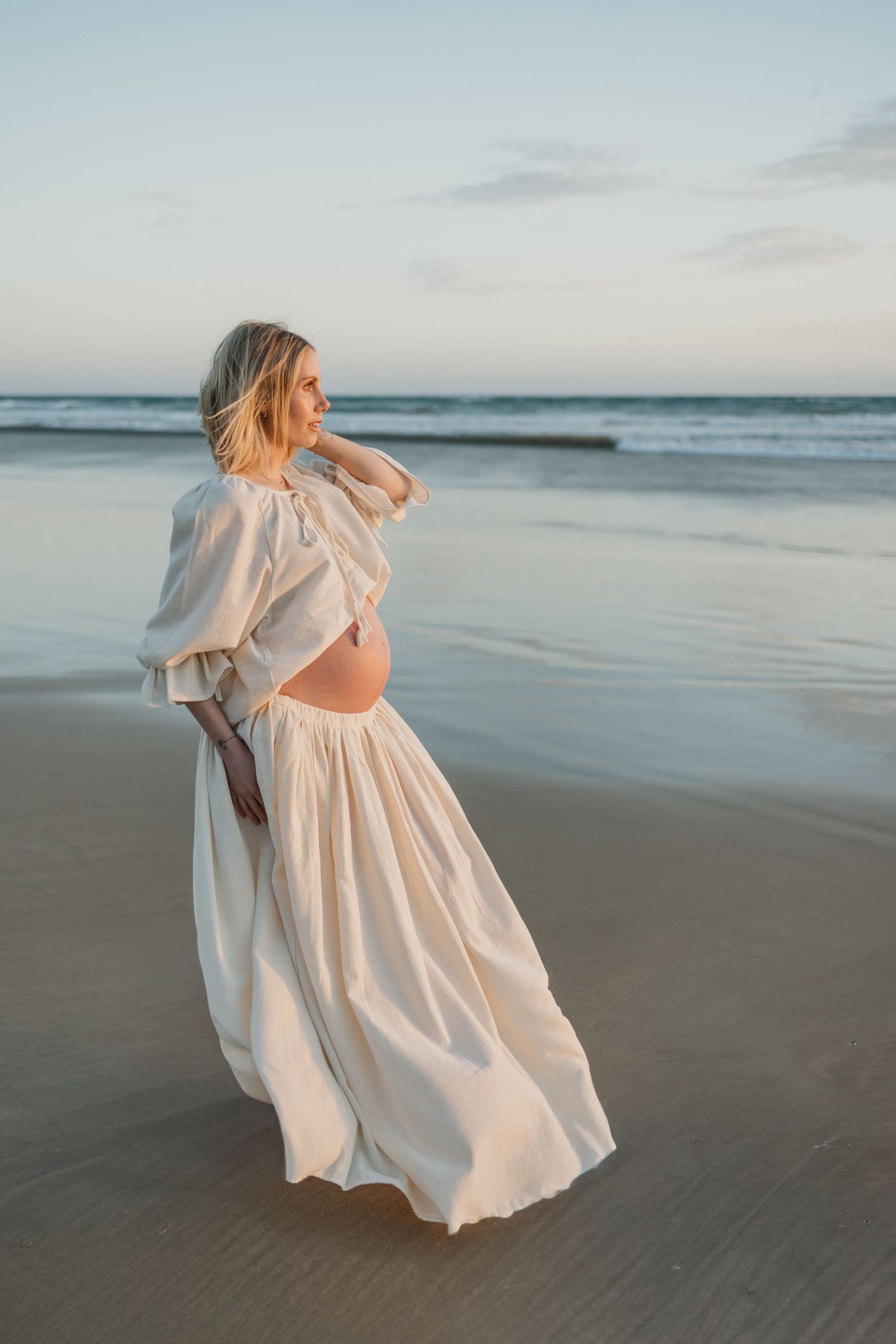 Pregnant woman in flowing white dress poses on beach at sunset with ocean waves behind her.