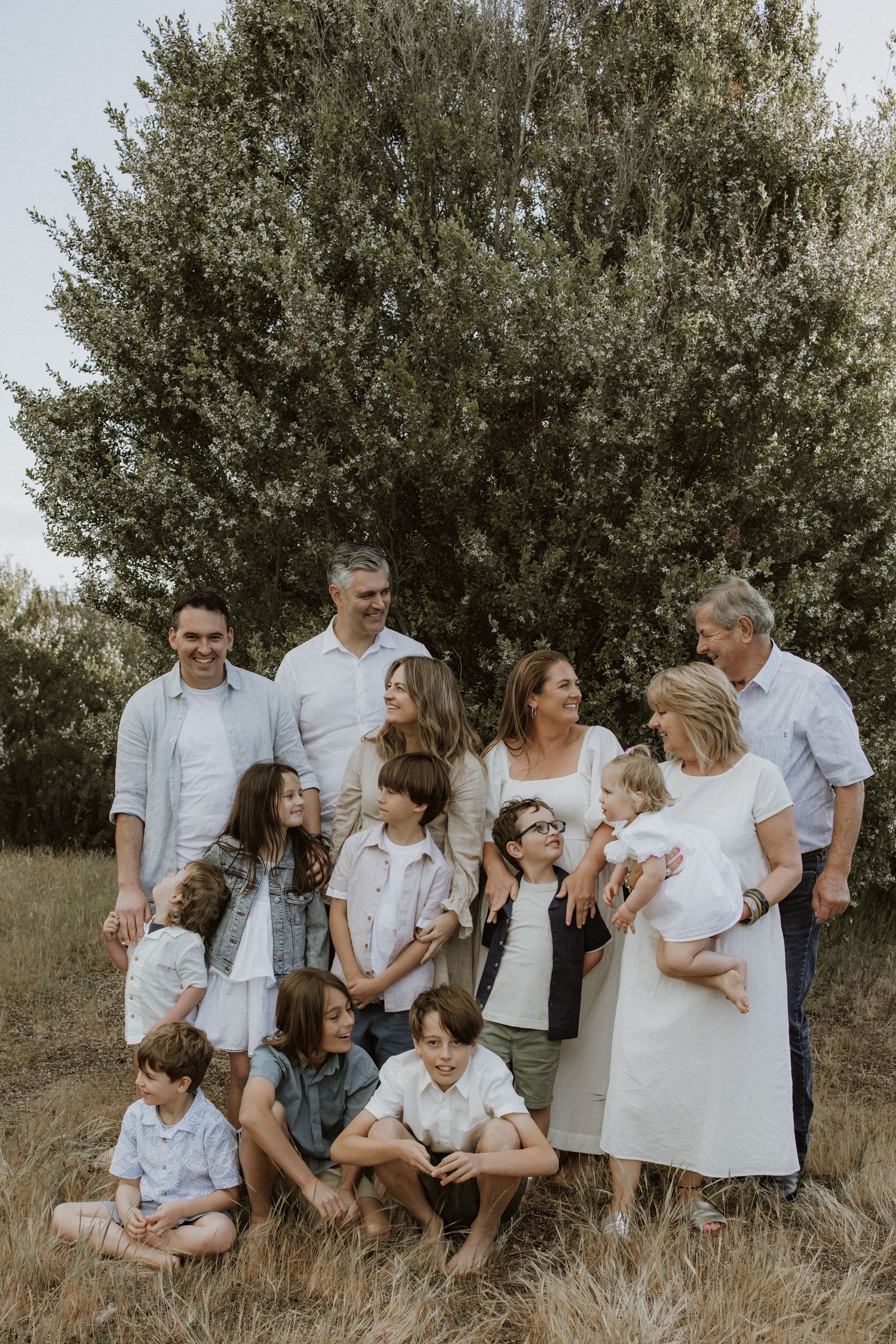 Large family group portrait outdoors in a field, posing together near a green tree on a sunny day.