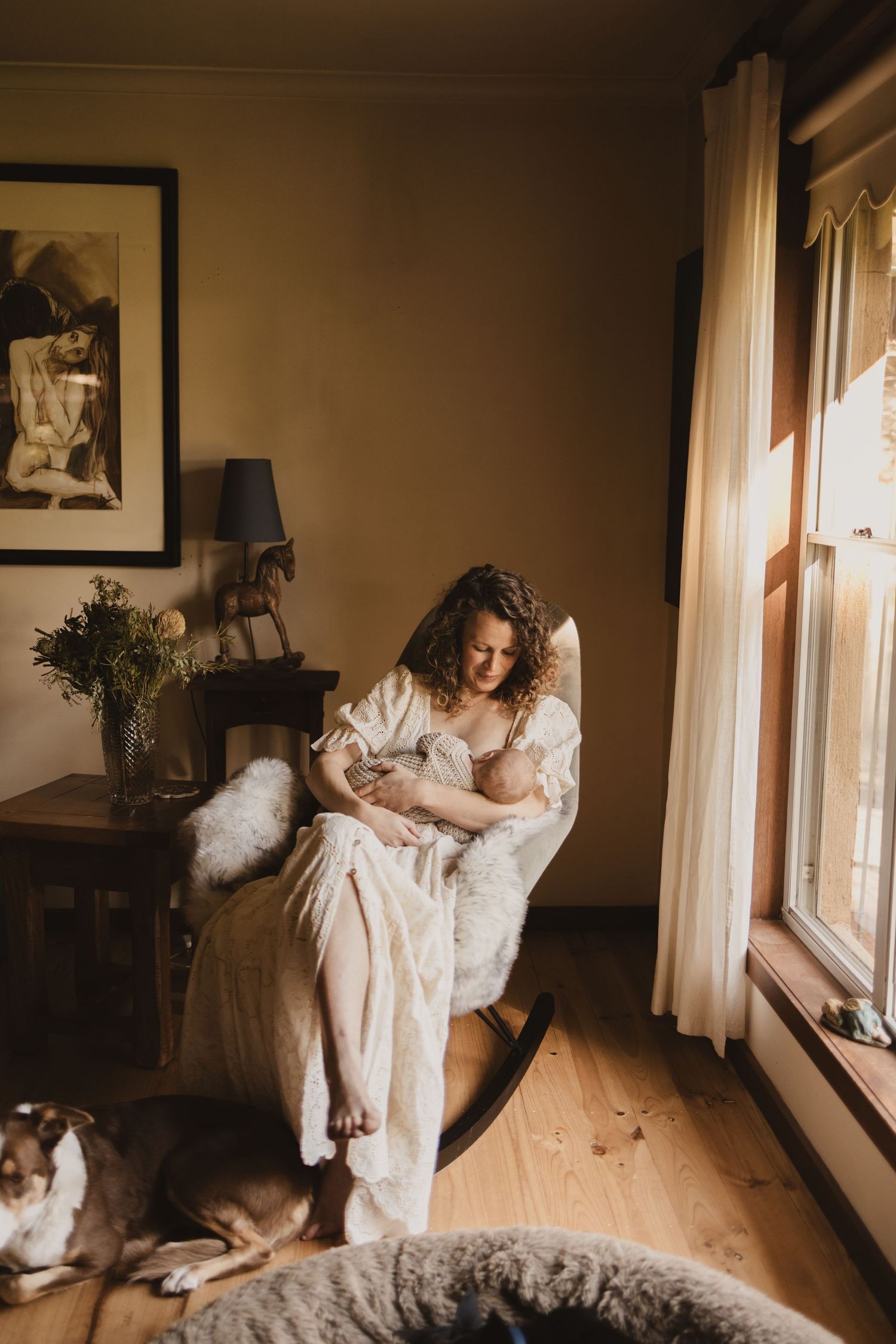 Mother breastfeeding newborn in rocking chair by sunlit window, with dog resting nearby on wooden floor.