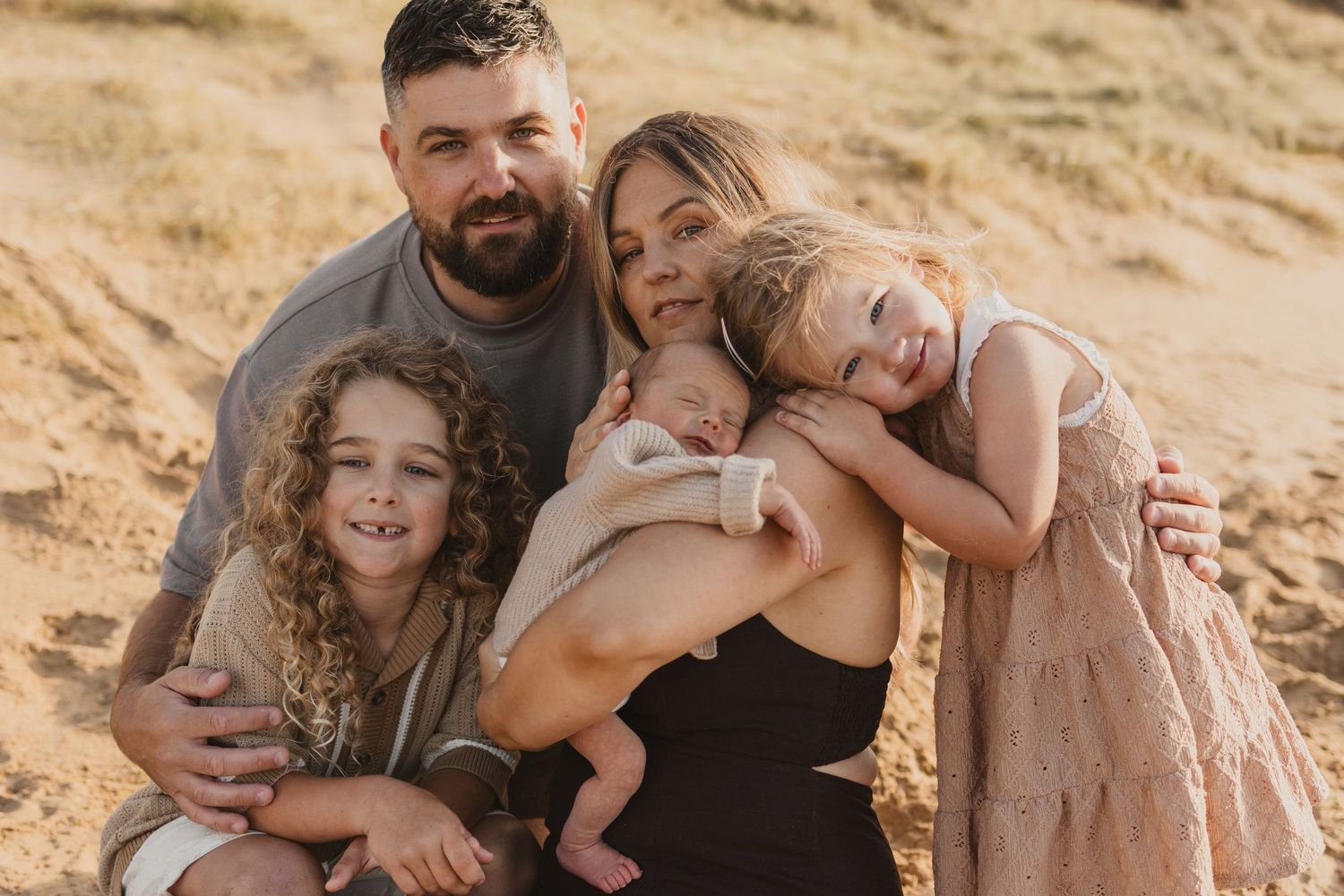 Happy family of five posing on sandy beach, parents holding newborn baby with two young children smiling outdoors.