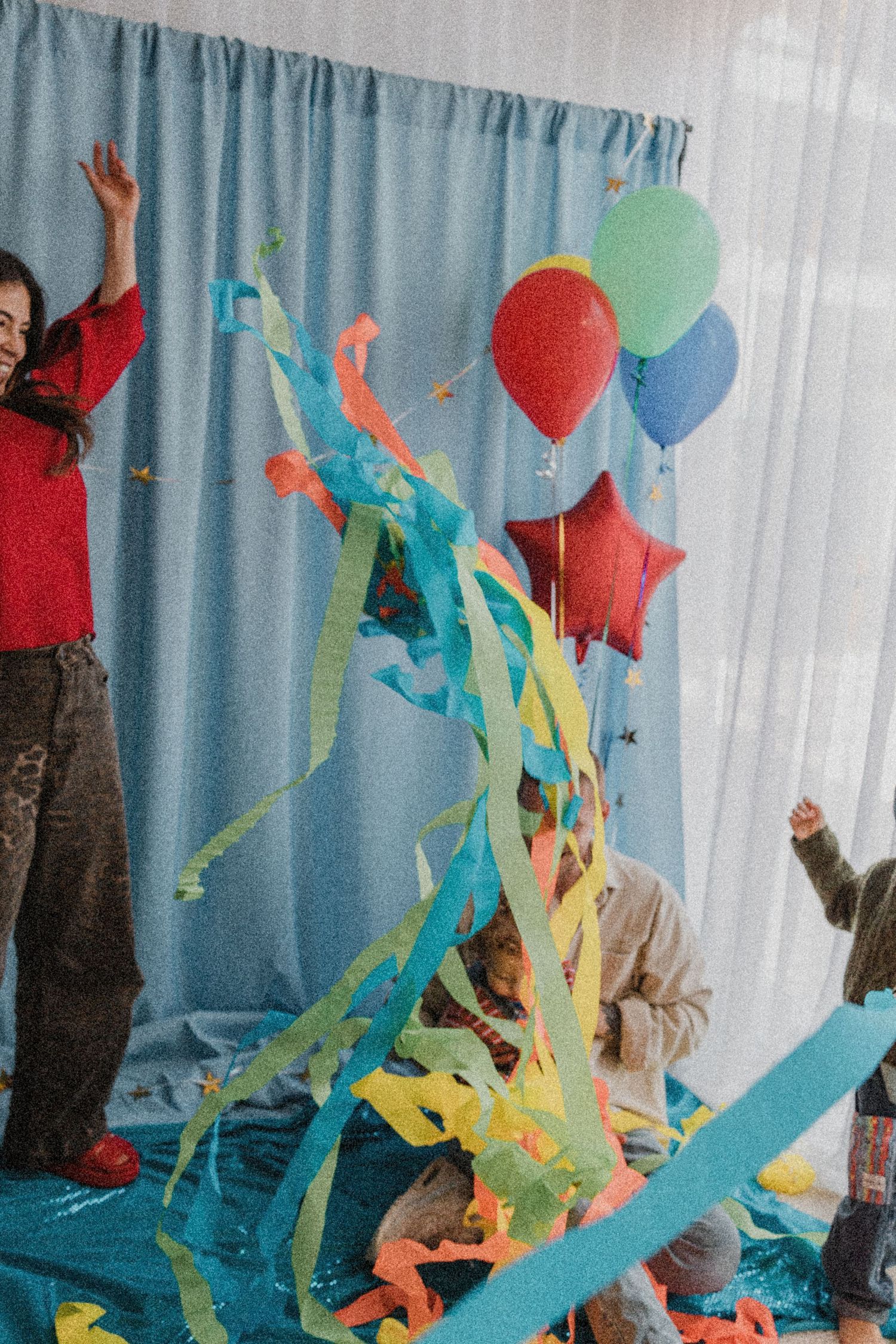 Children celebrate with colorful streamers, balloons, and confetti at a festive party with blue curtain backdrop.