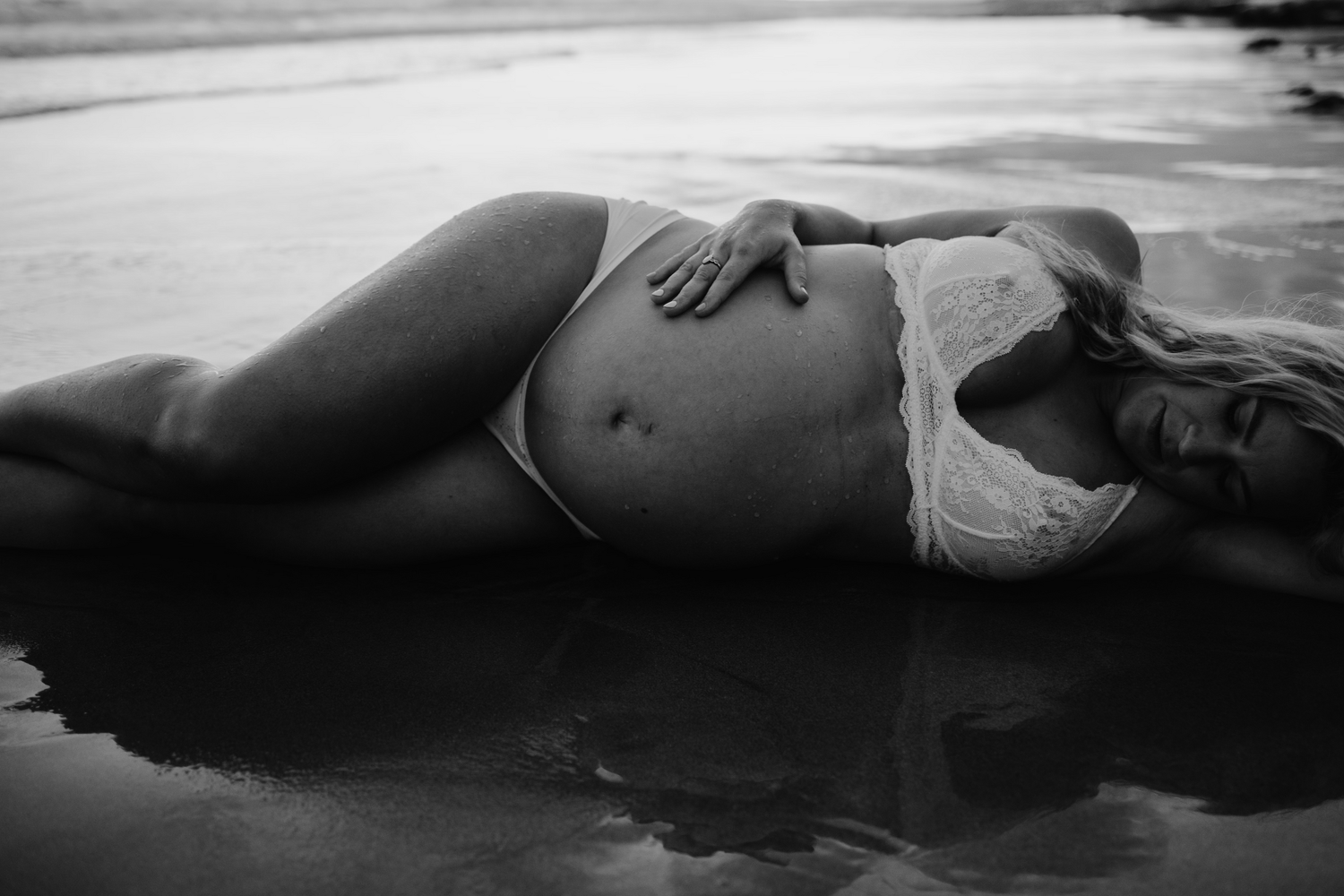 Black and white photo of a pregnant woman in lingerie lying on a beach, showcasing her baby bump.