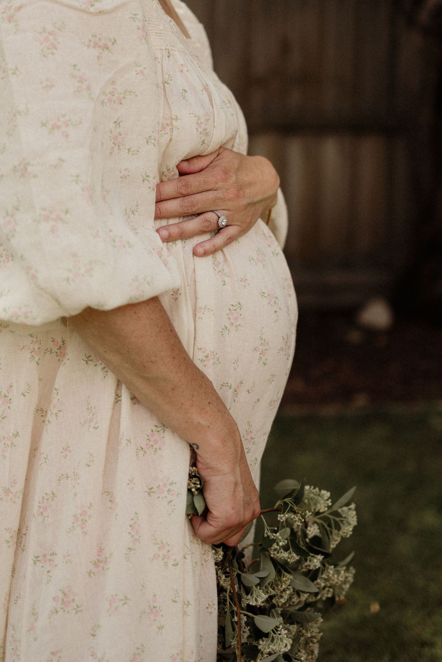 Pregnant woman in floral dress cradling baby bump, holding wildflowers, maternity photography outdoors.