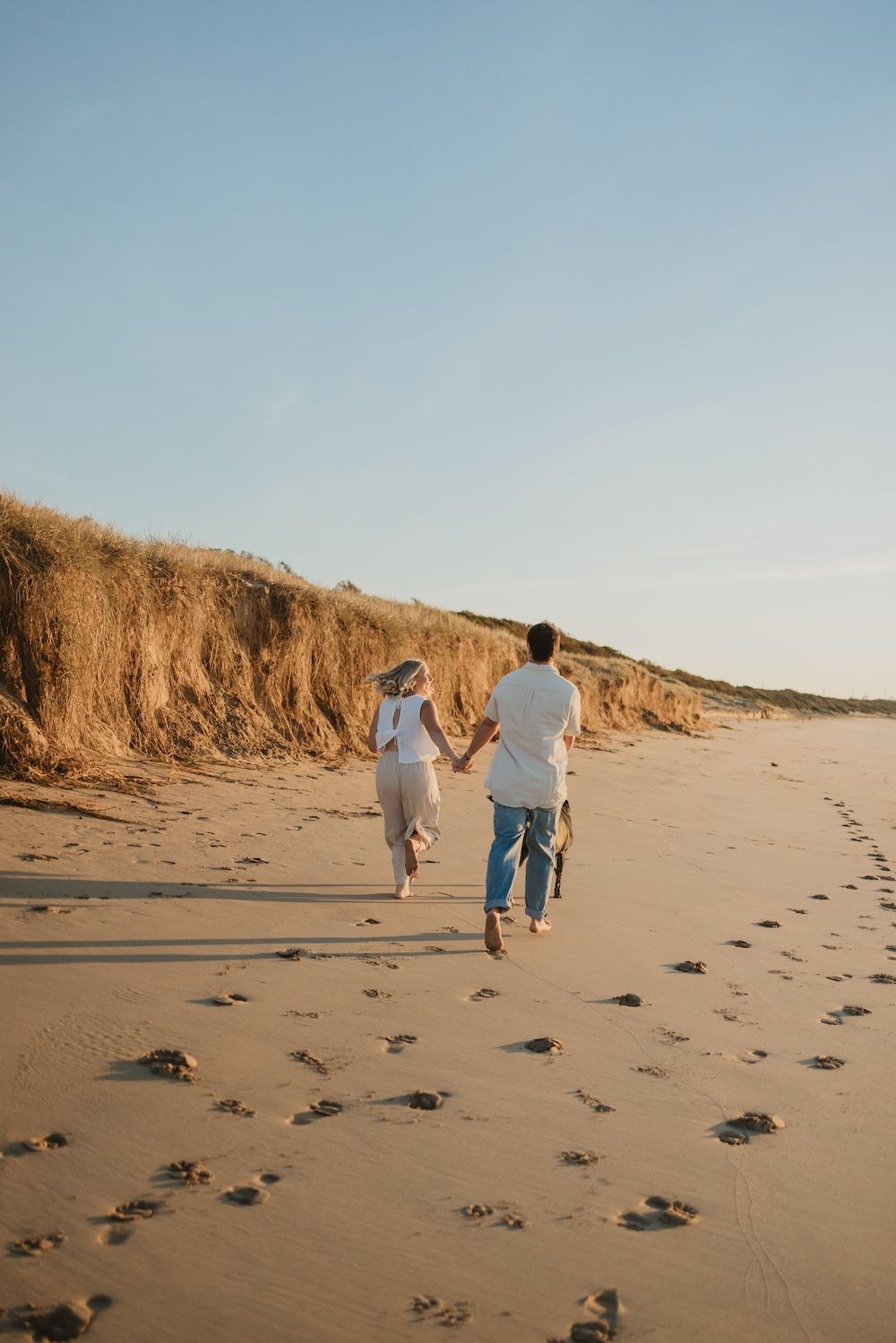 Couple holding hands running barefoot on sandy beach with golden dunes and clear blue sky at sunset.