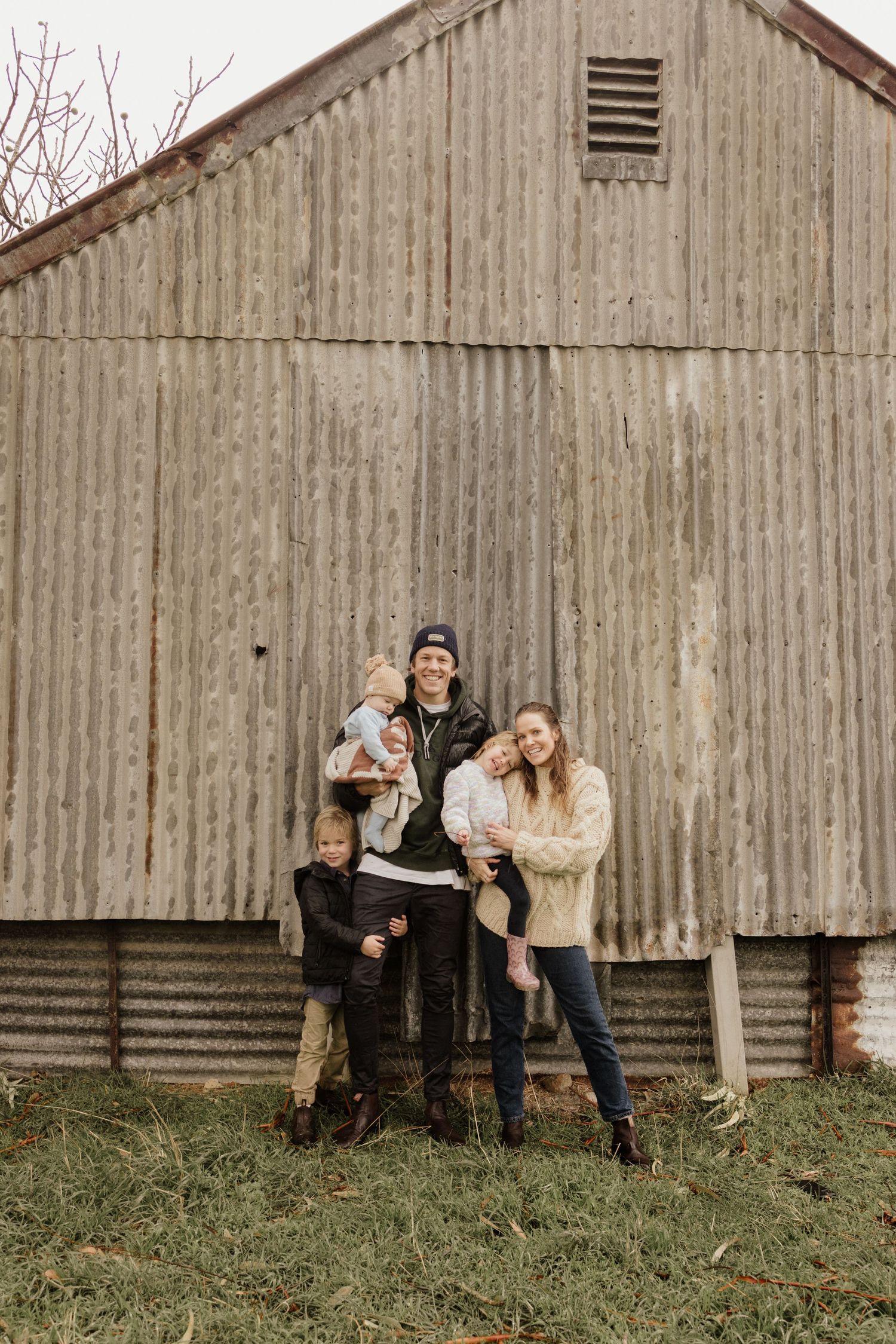 Family of four posing happily in front of a rustic corrugated metal barn on a farm.