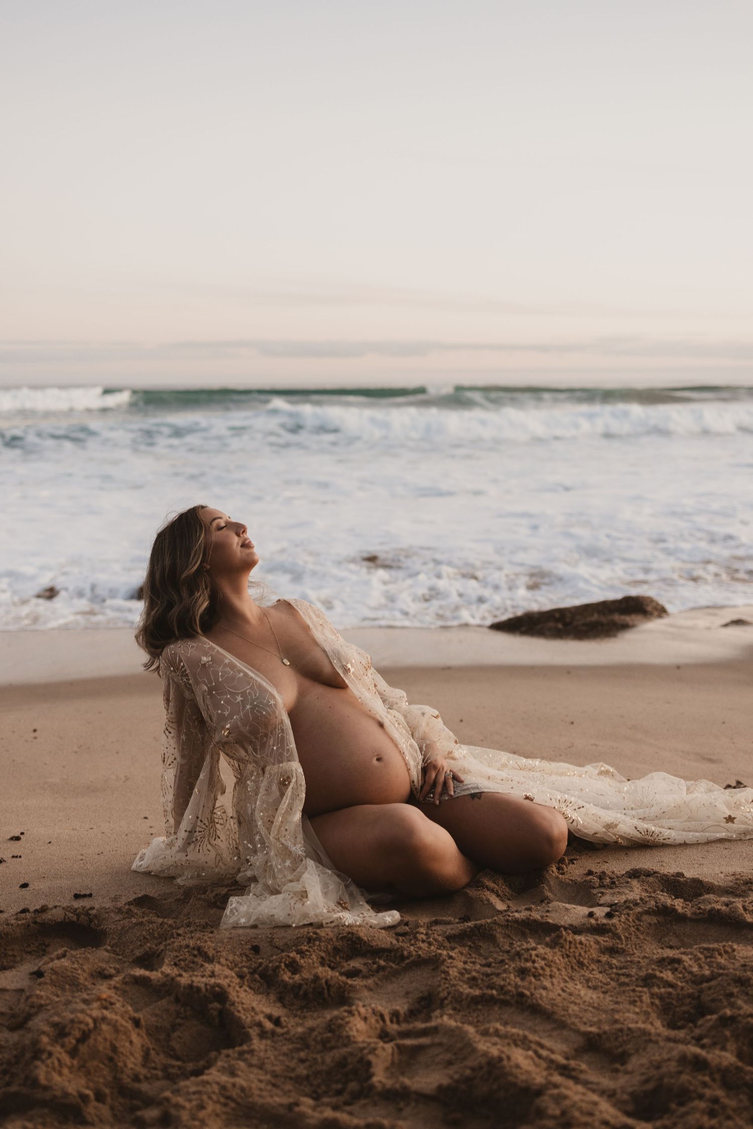 Pregnant woman in lace robe sitting on sandy beach at sunset with ocean waves in background.