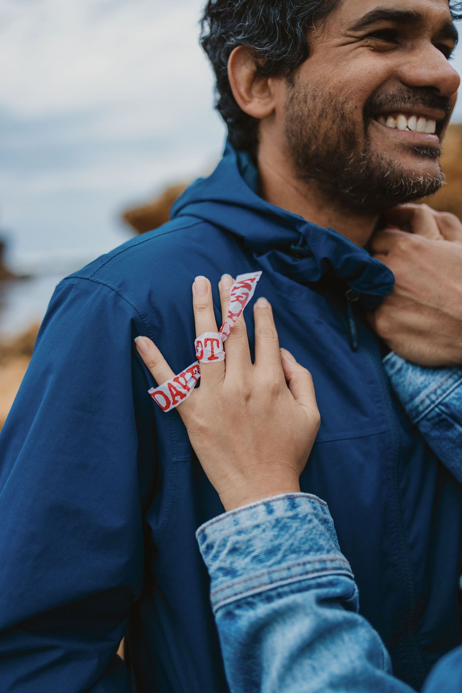 Smiling bearded man in blue jacket embraced from behind by person wearing festival wristband outdoors.