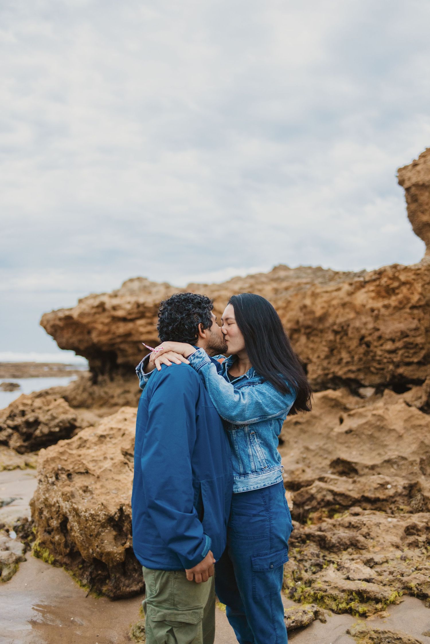 A couple sharing a romantic kiss on a rocky beach, embracing each other in blue outfits on a cloudy day.