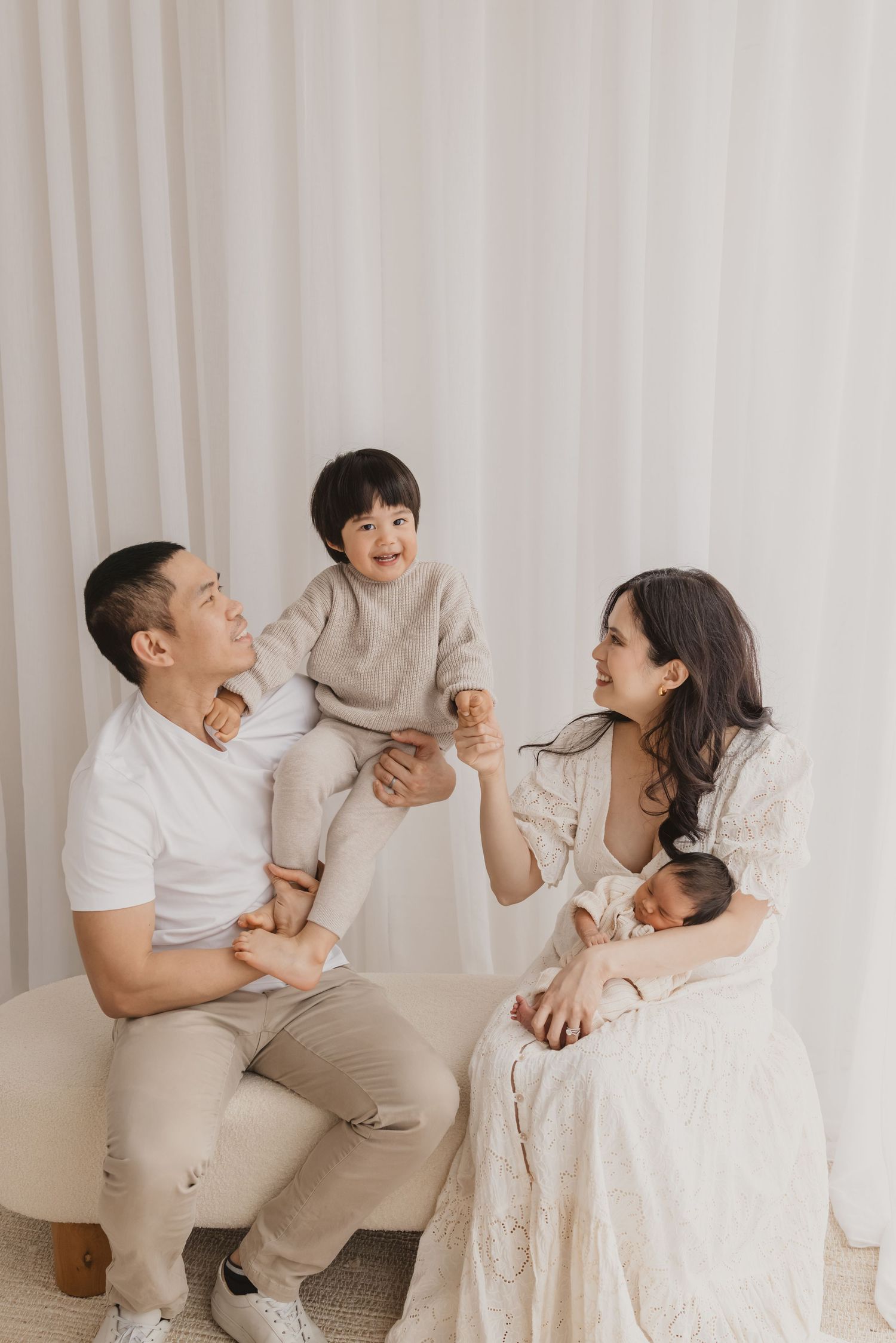A happy Asian family with a toddler and newborn baby poses together in a bright, neutral-toned studio setting.