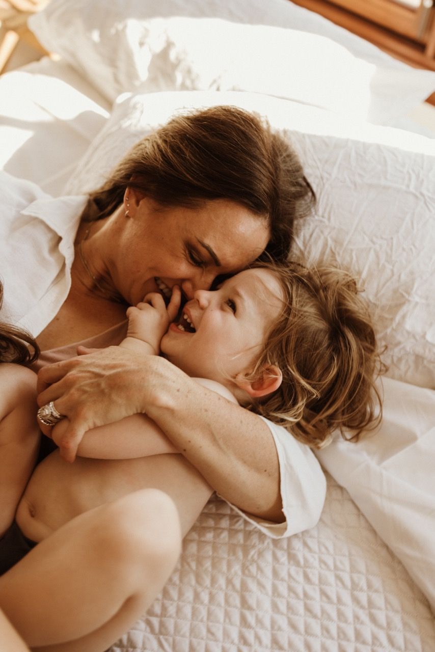 Tender moment shared between parent and child lying on white bedding in morning light.