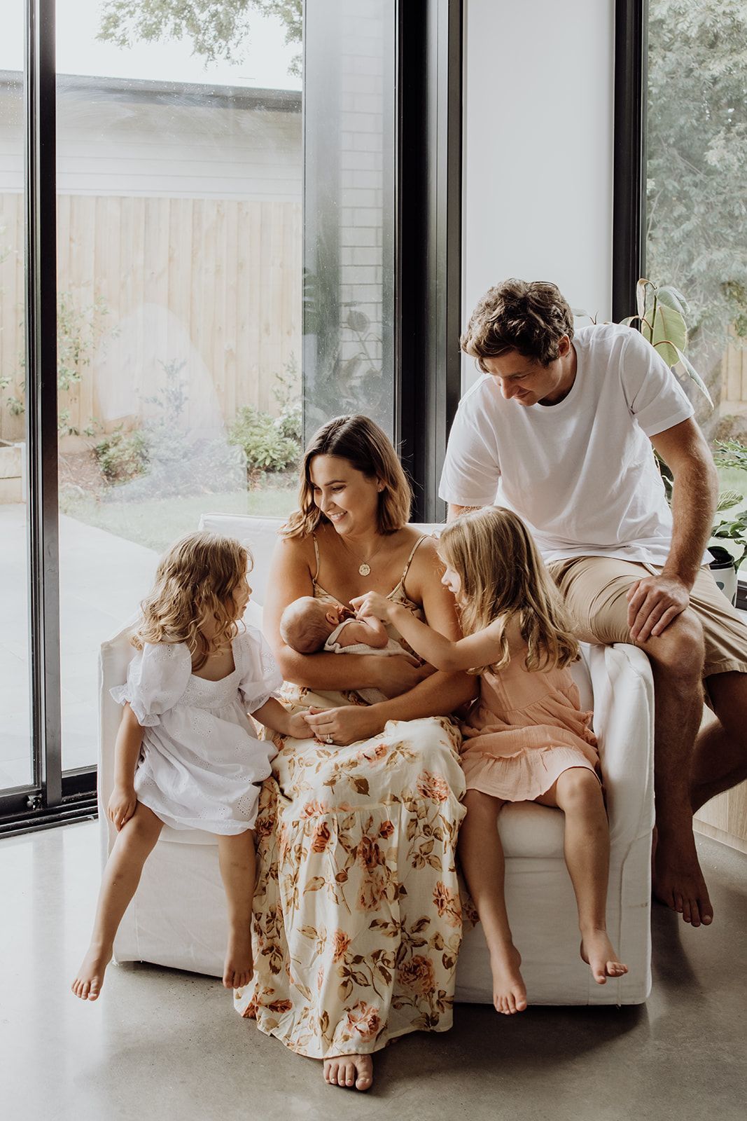 Family of five with newborn baby gathered on white sofa near large windows in modern bright home.