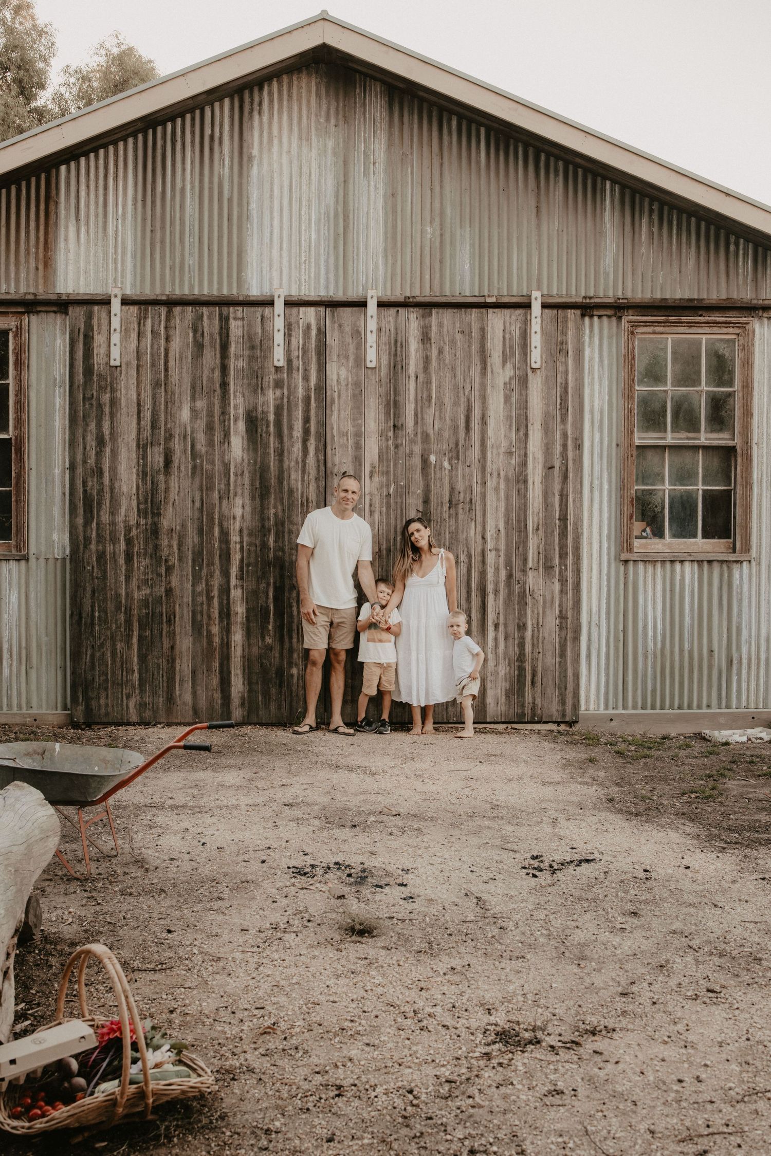 A family of three poses in front of a rustic corrugated metal barn on a dirt lot.