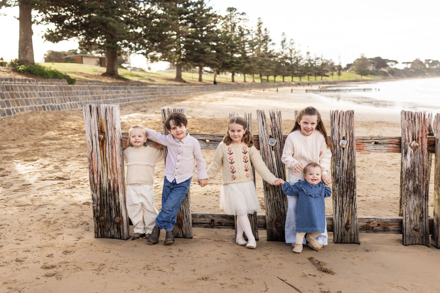Five young children hold hands and smile on a sandy beach surrounded by rustic wooden posts with trees in the background.